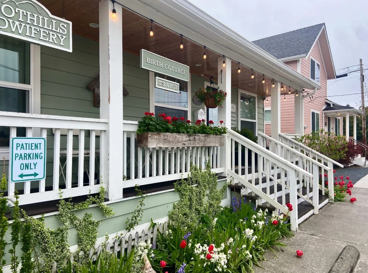 A quaint green porch with white railings and stairs. There are plants and flowers along the base and a wooden flower box with red flowers. Hanging string lights are above the porch. The house has a sign that says 'Birth Cottage' and 'It's a Boy,' indicating a birth announcement. A sign on the railing reads 'Patient Parking Only.'