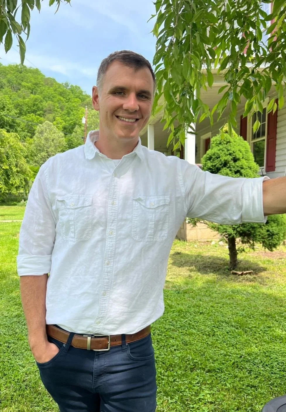 A smiling man with short brown hair wearing a white button-up shirt with rolled-up sleeves, standing outdoors on a bright day in a lush green yard with trees and a house in the background.