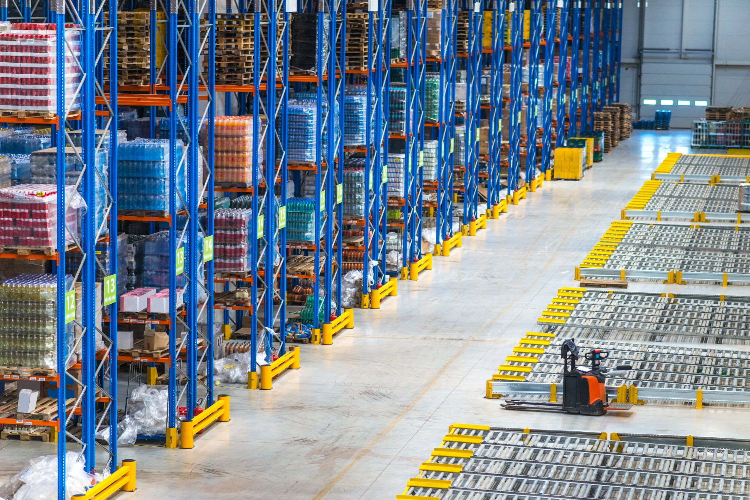 A large warehouse with tall blue and orange shelving racks filled with various goods. The floor has yellow safety barriers and some empty racks or containers.