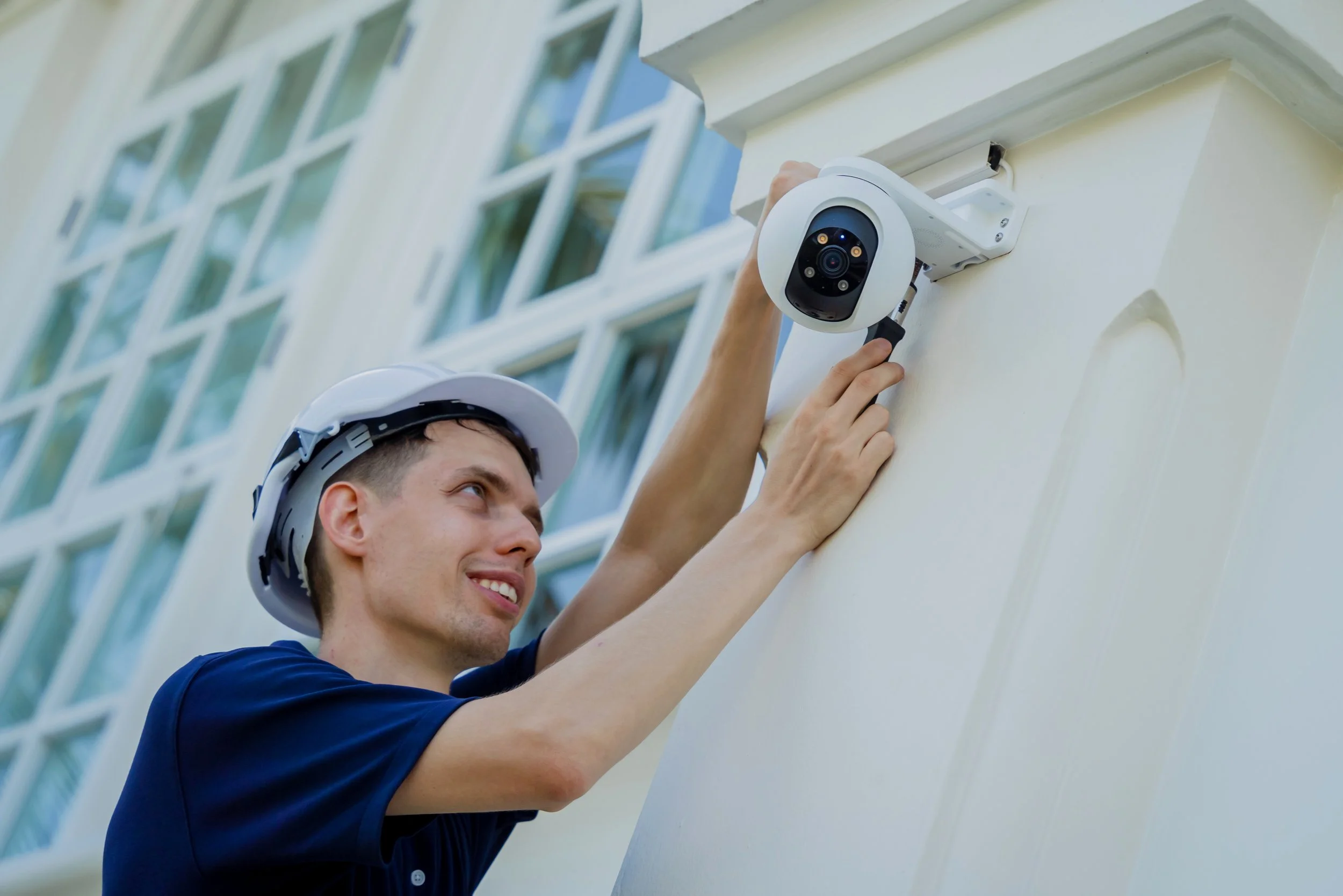A man wearing a white safety helmet installs or inspects a security camera on an exterior wall of a building with large windows.