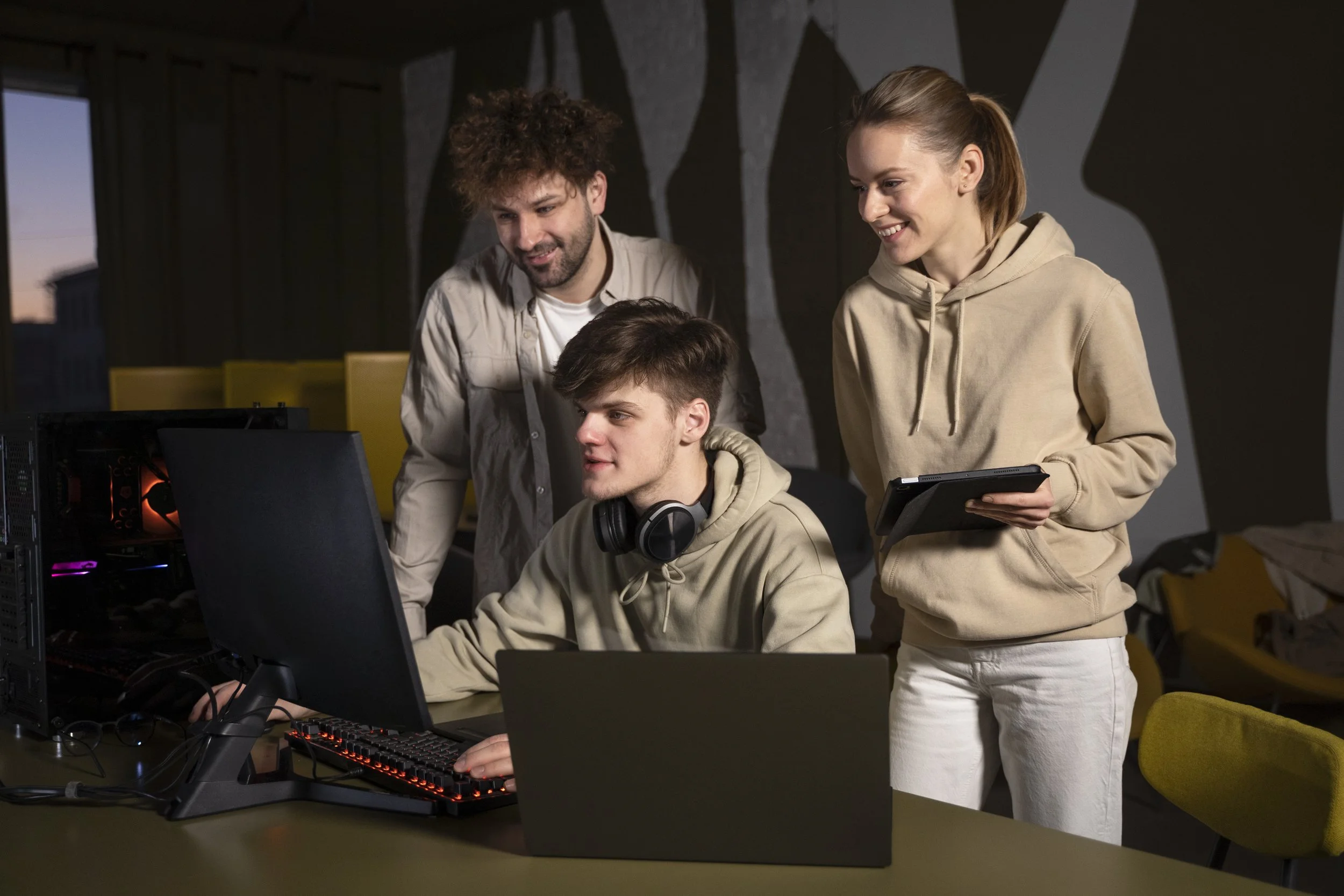 Three young people gathered around a computer in a gaming setup, with one seated at the computer wearing headphones, and two standing beside him, one holding a tablet, smiling and engaging with each other.