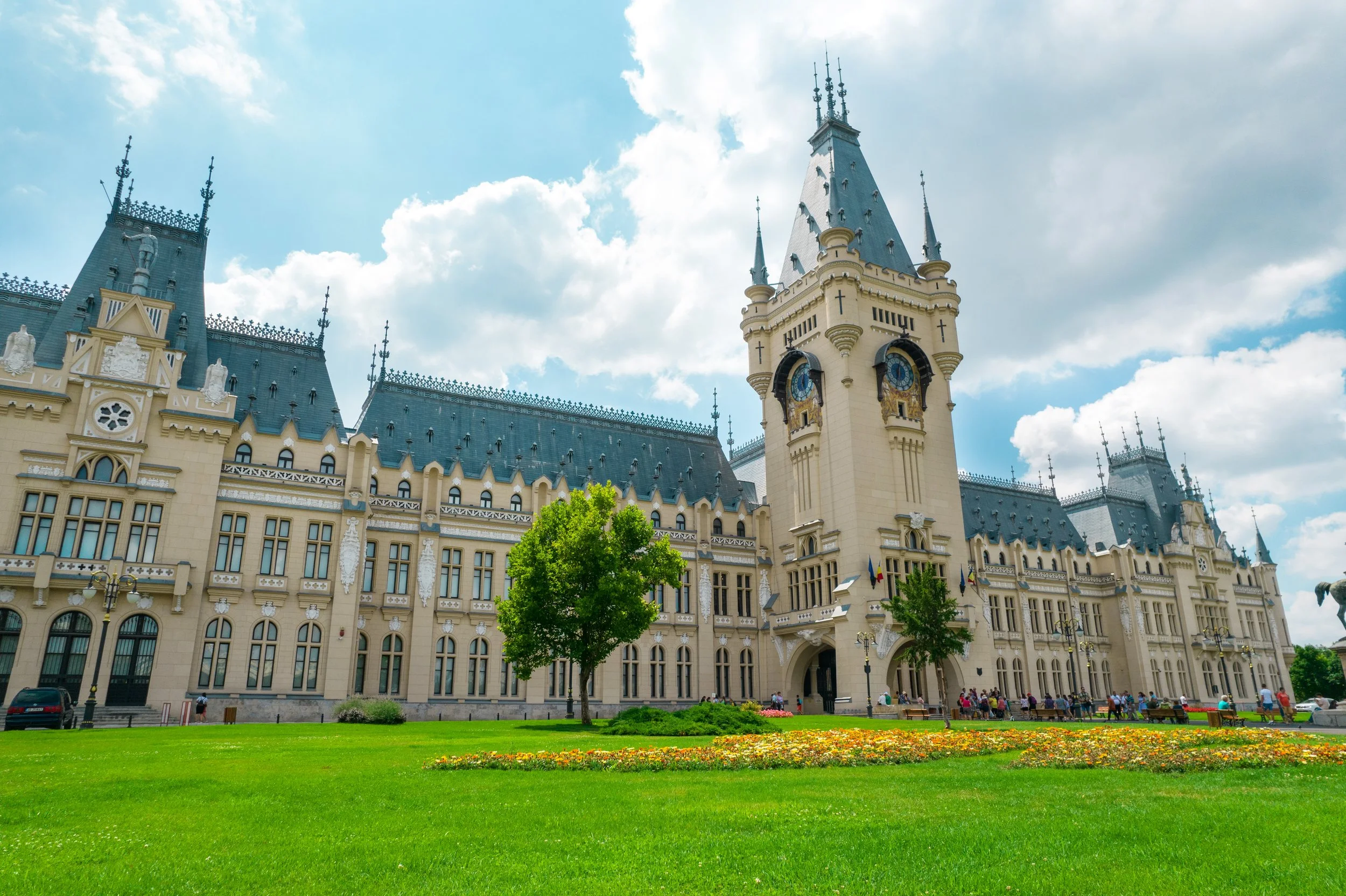 A large, castle-like building with a tall clock tower, multiple turrets, and ornate architecture, set against a partly cloudy sky with a green lawn and flowers in the foreground.