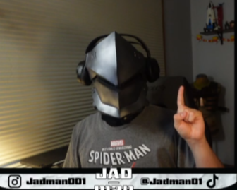 Person wearing a Genji helmet and headphones, pointing upward, in a room with blinds and shelves.