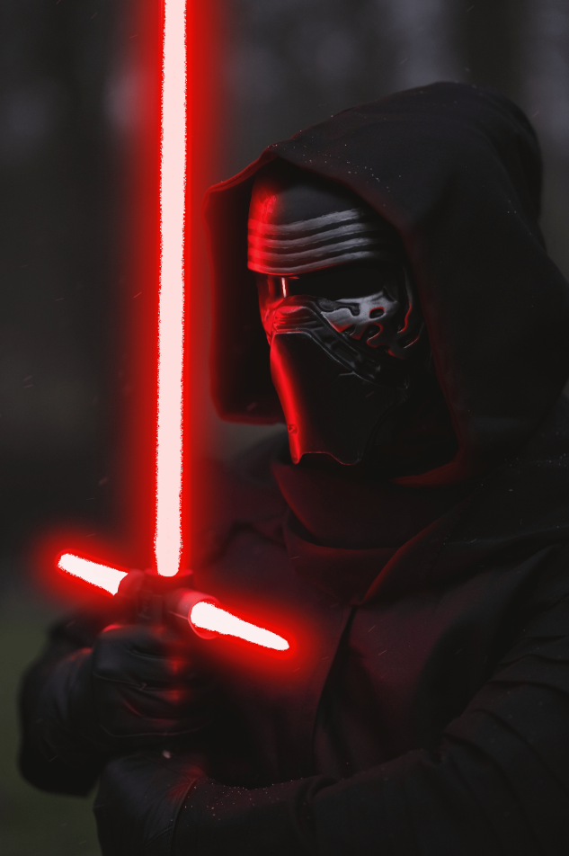 A person wearing a black hooded cloak and a shiny black mask holding a red glowing lightsaber, with a blurred dark forest background.