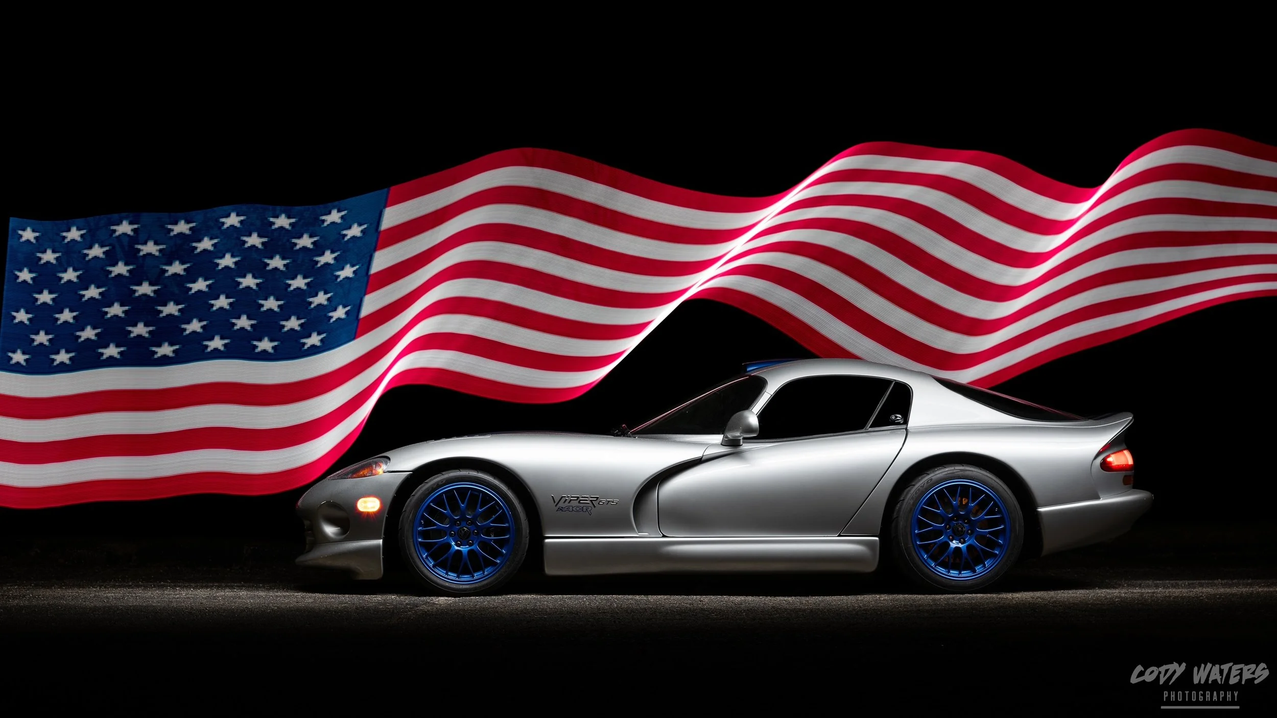 A silver Dodge Viper with blue rims parked against a black background with a waving American flag overhead.