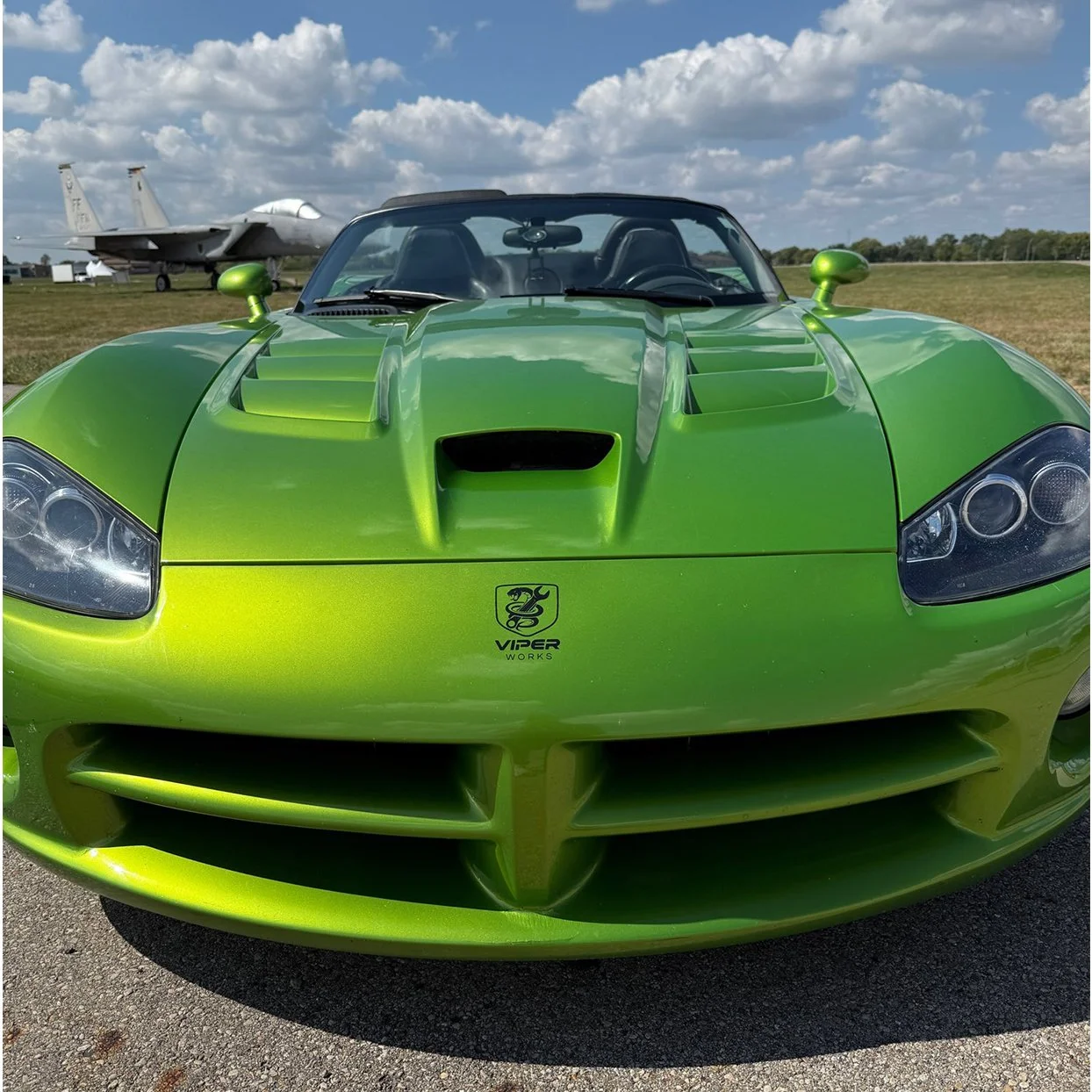A bright green Dodge Viper with a sleek, aerodynamic design and a prominent front grille, parked outdoors under a partly cloudy sky with a fighter jet airplane in the background.
