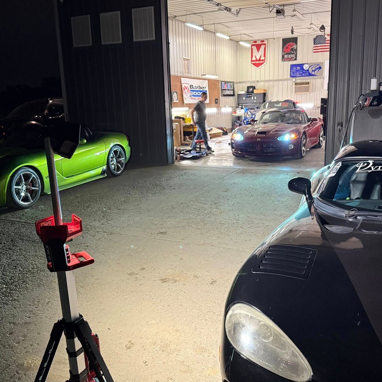 Inside a garage with cars, including a black Dodge Viper on the right, a purple Dodge Viper in the center, and a green Dodge Viper on the left. A person is working near the purple car, and tools and banners are hanging on the walls.