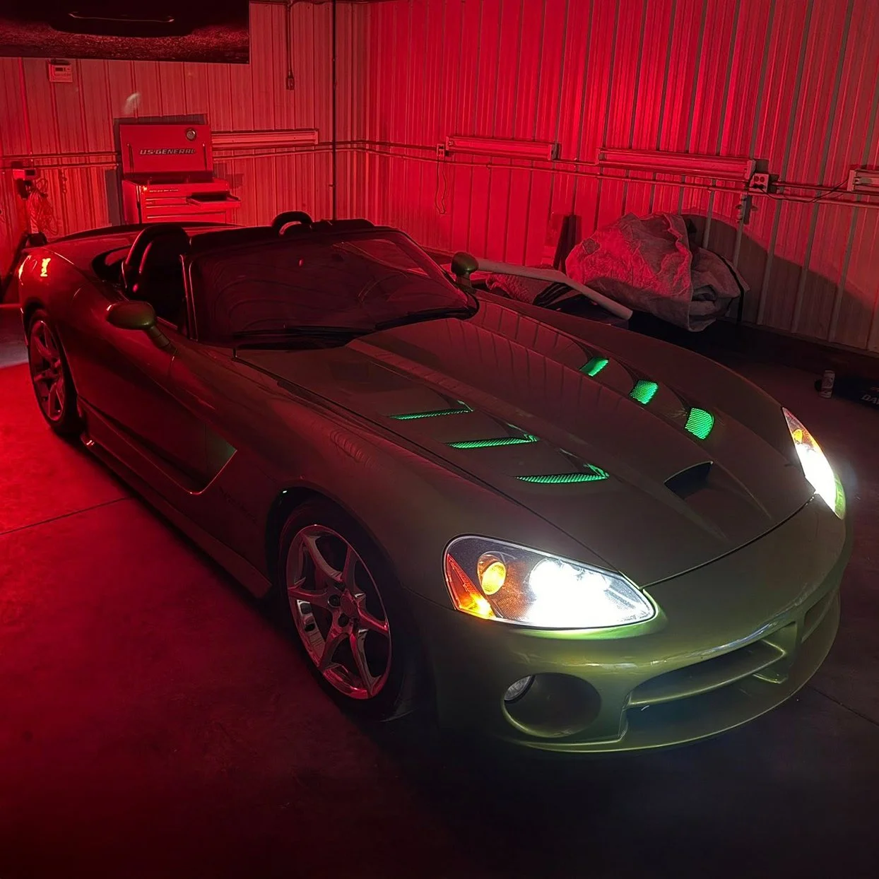 A Dodge Viper with green and white headlights parked inside a garage with red lighting, and a workbench with a cloth and tools.