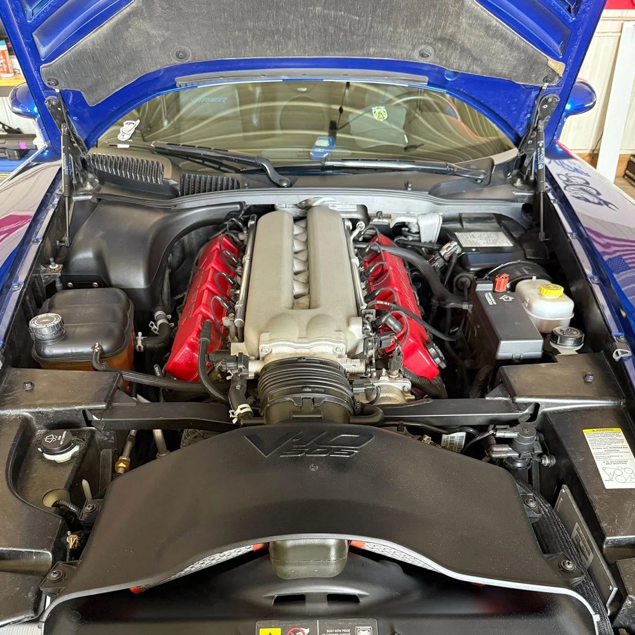 Engine bay of a blue Dodge Viper with a prominent V10 engine, featuring red valve covers, with various hoses, reservoirs, and components visible.