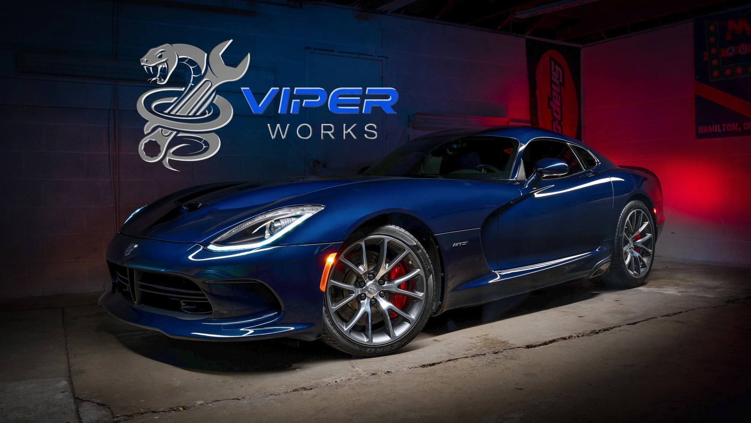 A blue Dodge Viper parked inside a garage with a Viper Works sign and logo on the wall, illuminated with red and blue lighting.