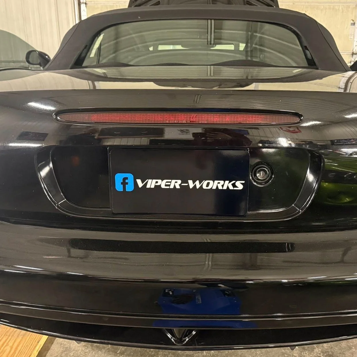 Rear view of a black Dodge Viper with a custom license plate that reads 'VIPER-WORKS' and a Facebook logo. The car is parked inside a garage.