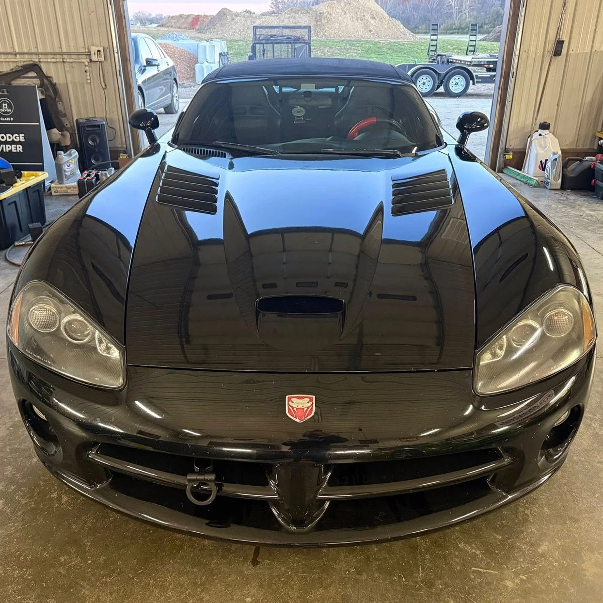 Black Dodge Viper with an emblem on the front, parked inside a garage with various tools and equipment around, and outdoor landscape visible through the open garage door.