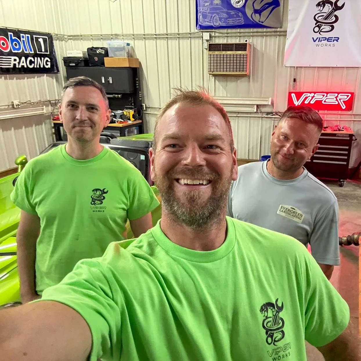 Three men smiling at the camera inside a garage or workshop with automotive signs and tools. Two men are wearing bright green shirts with a snake logo and the text 'Viper Works.' The third man is wearing a gray shirt with a logo and text 'Evergreen Landscaping.'