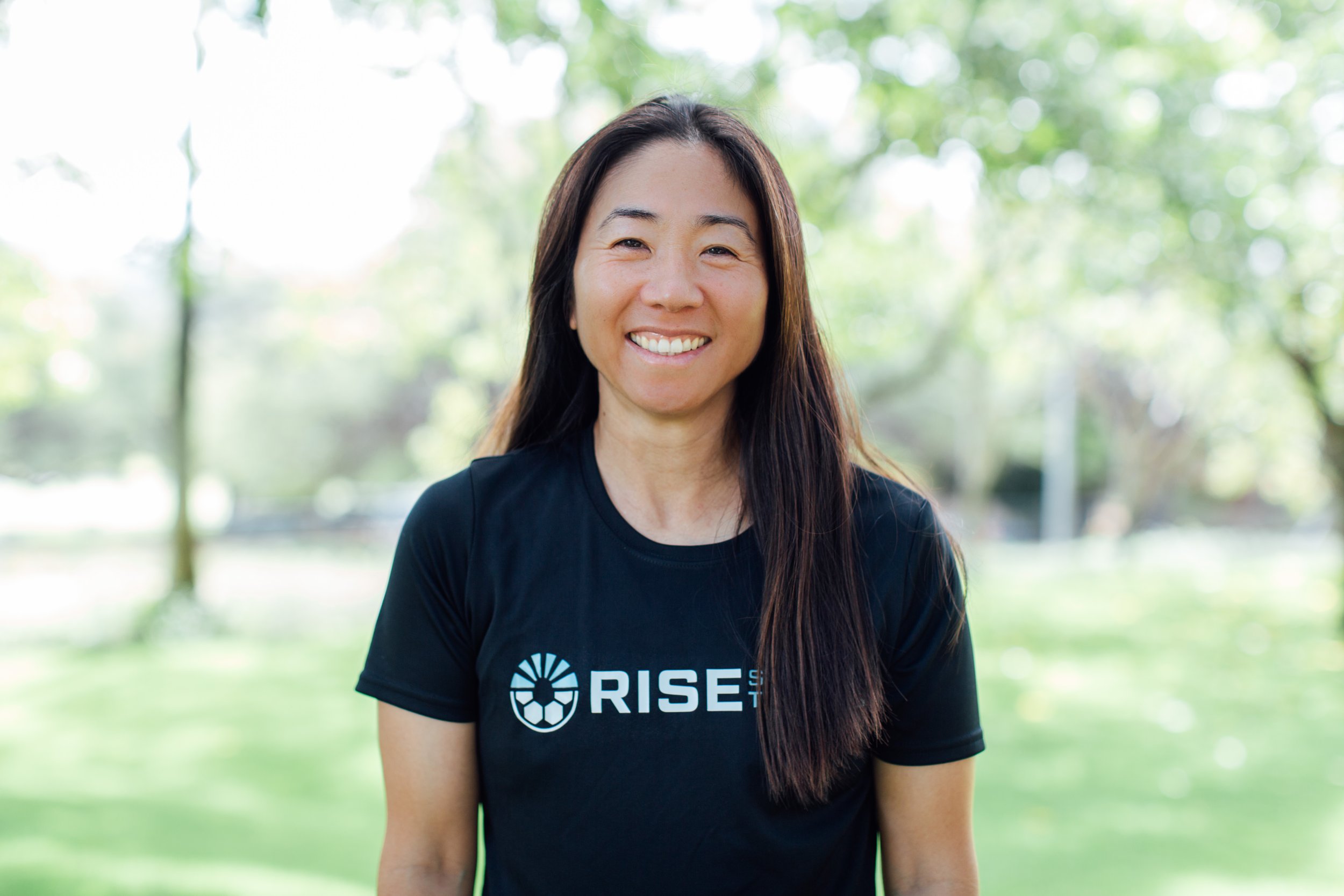 Smiling woman in a black shirt with 'RISE' logo standing outdoors in a park with green trees in the background.