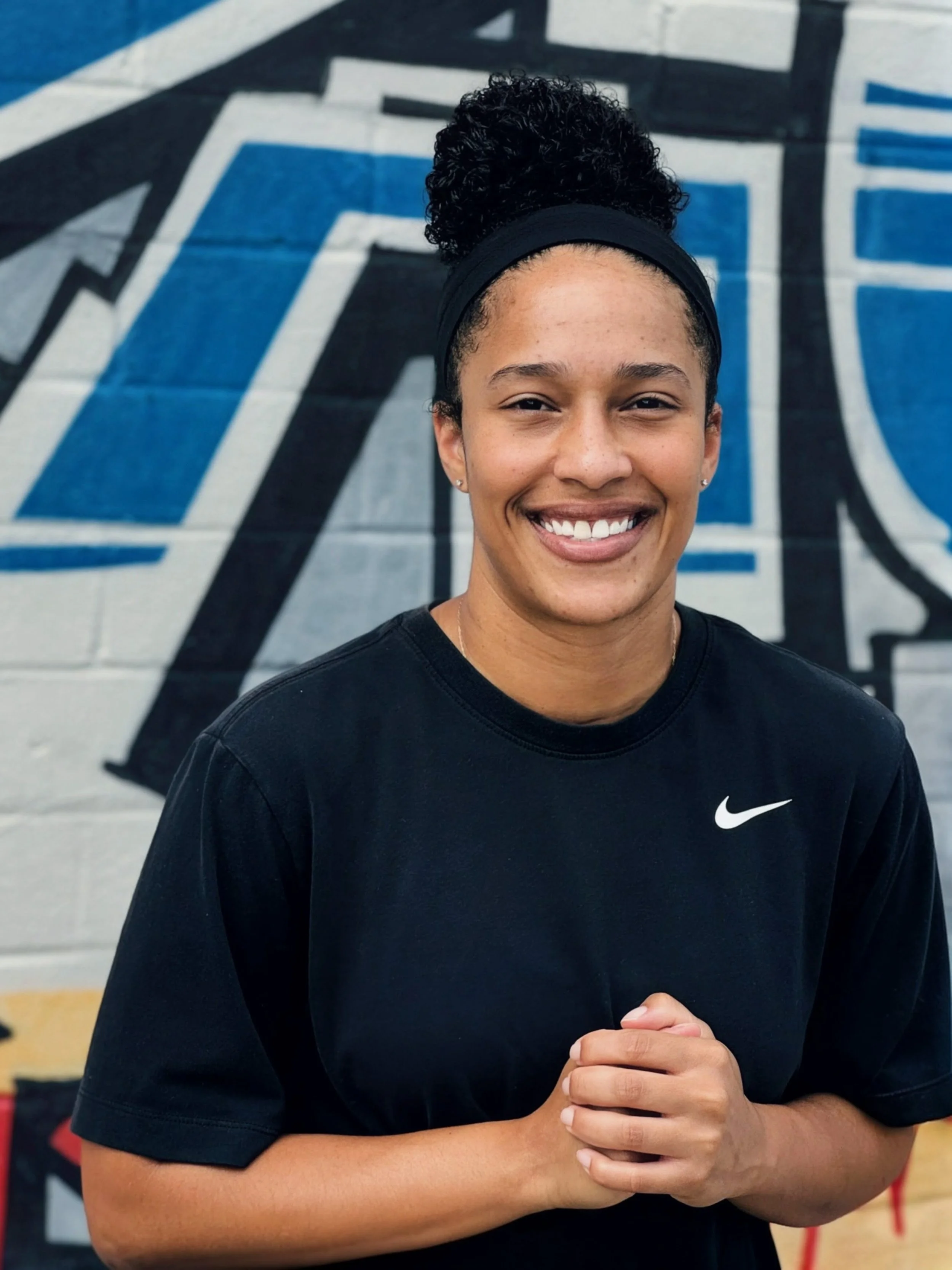 A young woman with curly hair tied back with a headband, smiling, wearing a black Nike shirt, standing in front of a colorful graffiti wall.