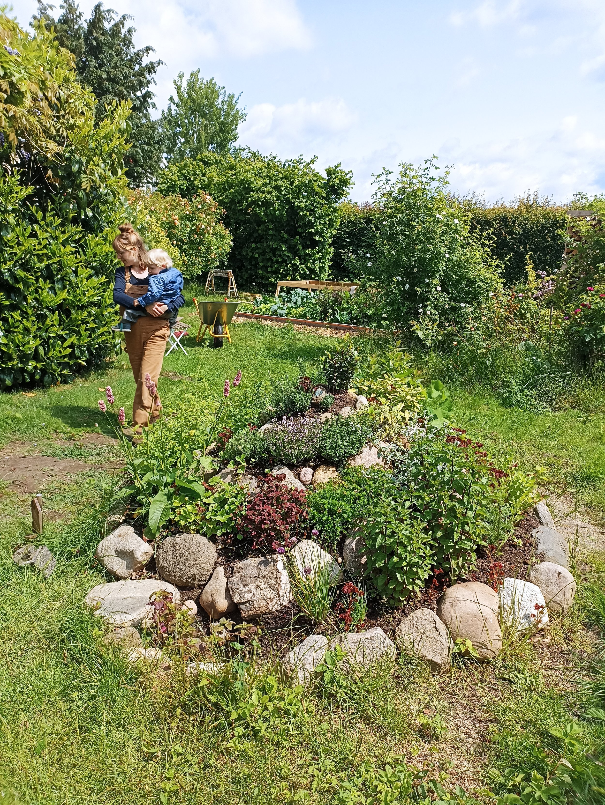 En kvinde går igennem en skovhave med et barn på armen en varm sommerdag. Kvinden går forbi en nyanlagt urtespiral med spiselige urter i blomst. I urtespiralen er der en summen af liv fra insekter og padder.
