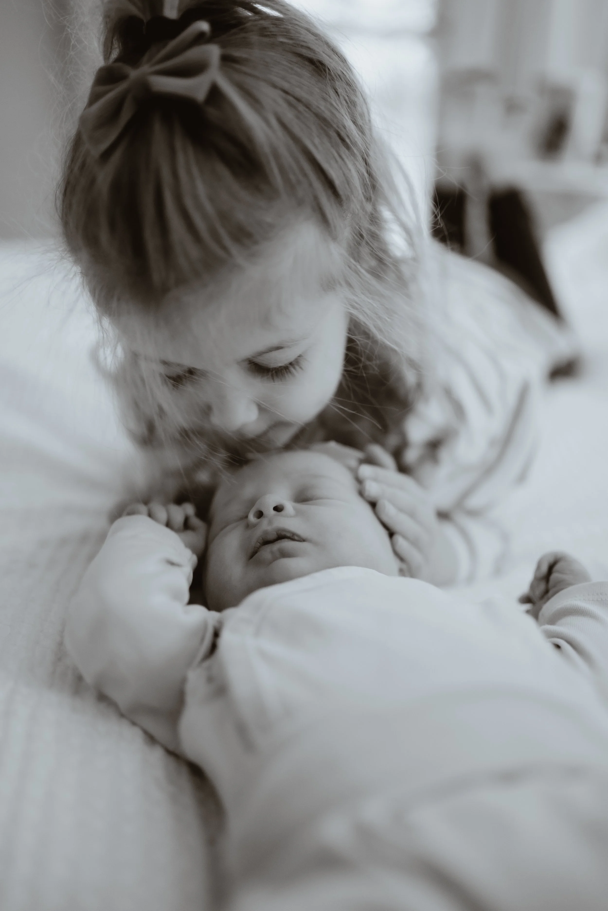 A young girl leaning over and kissing a sleeping baby on the forehead in a softly lit room. Fredonia, NY