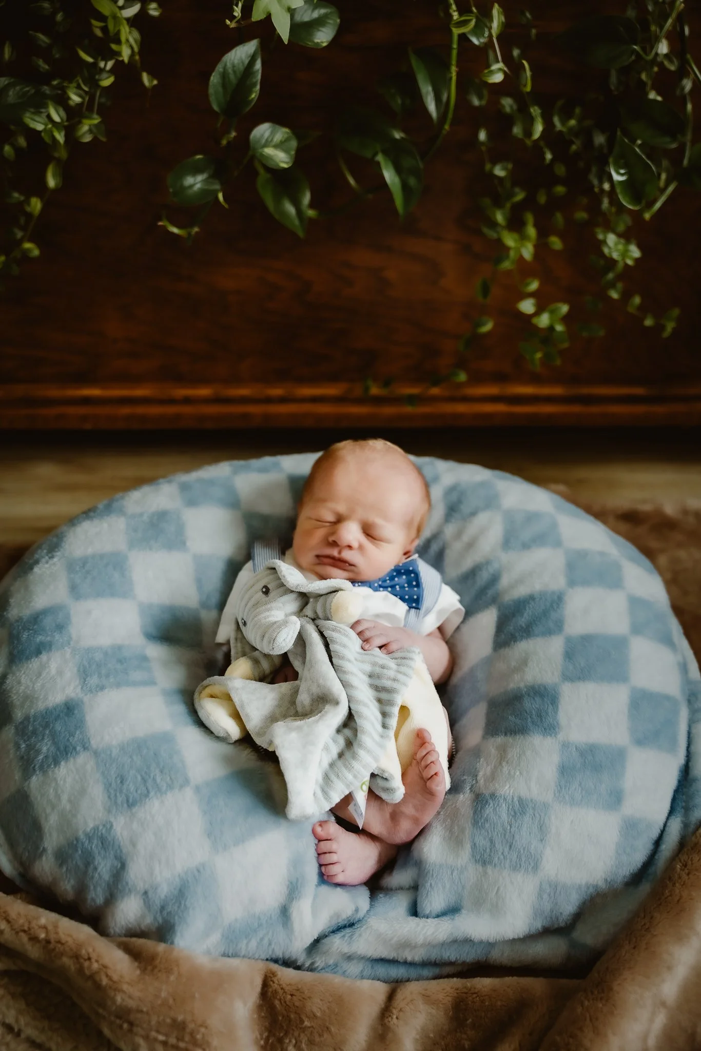A newborn baby with a pacifier laying on his back in a cushioned nest, wearing a white shirt with a blue bow tie and beige shorts, on a soft blanket with a checkered pattern.