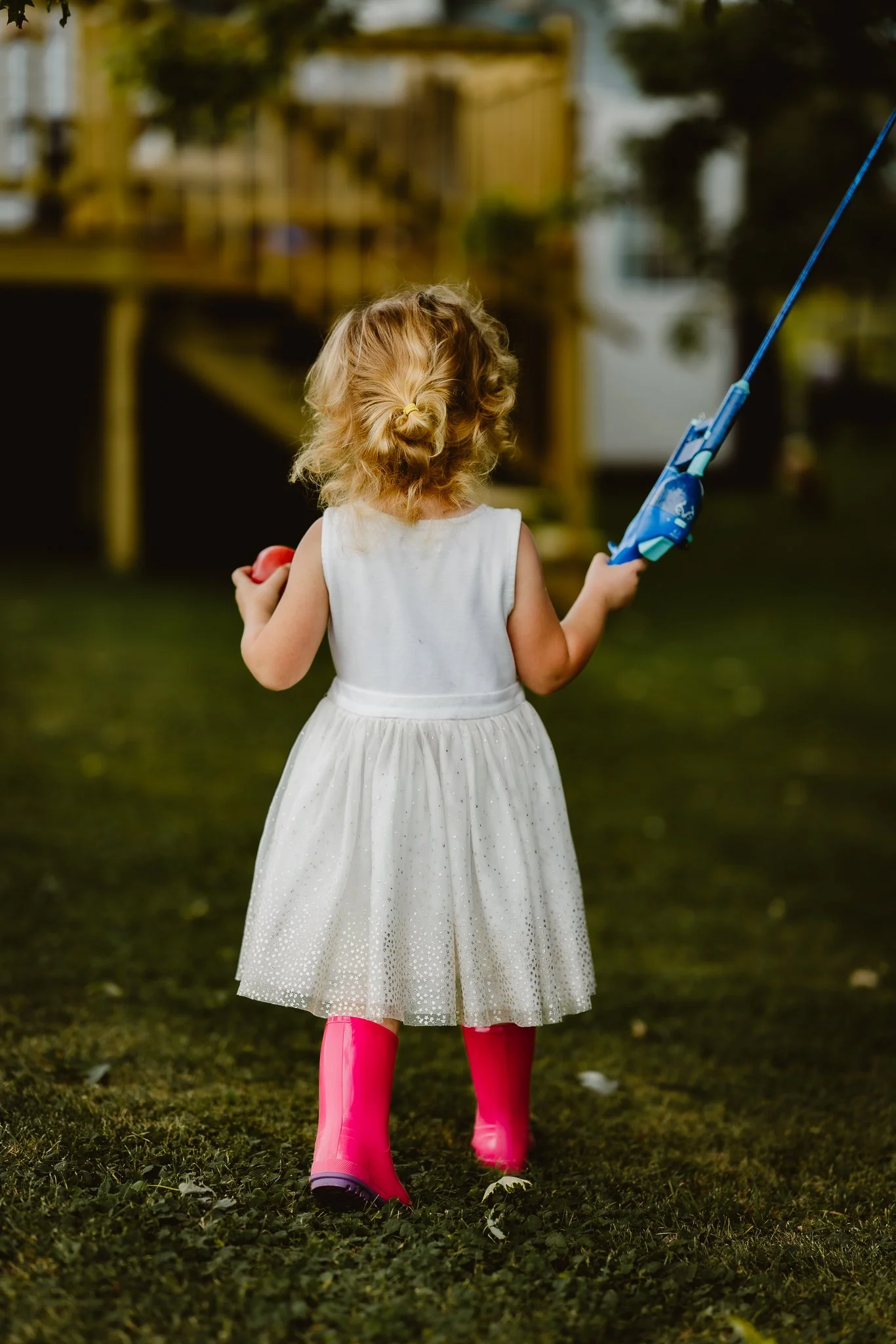 A young girl wearing a white dress and pink rain boots is in a backyard, holding a toy gun and a ball, with a wooden play structure and trees in the background.