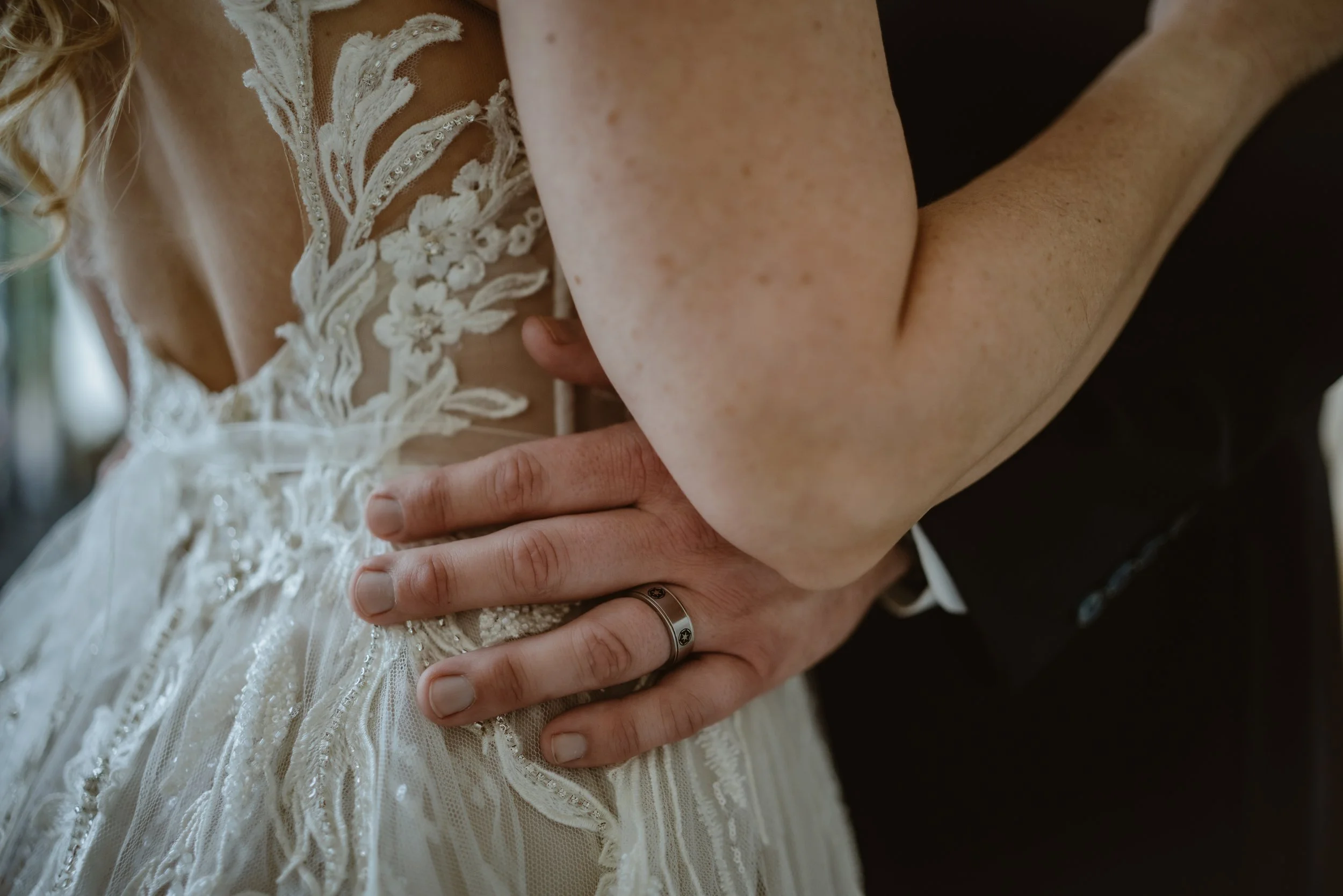 Close-up of a couple's hands, with the bride wearing a wedding dress with lace details, and the groom wearing a ring, on their wedding day.