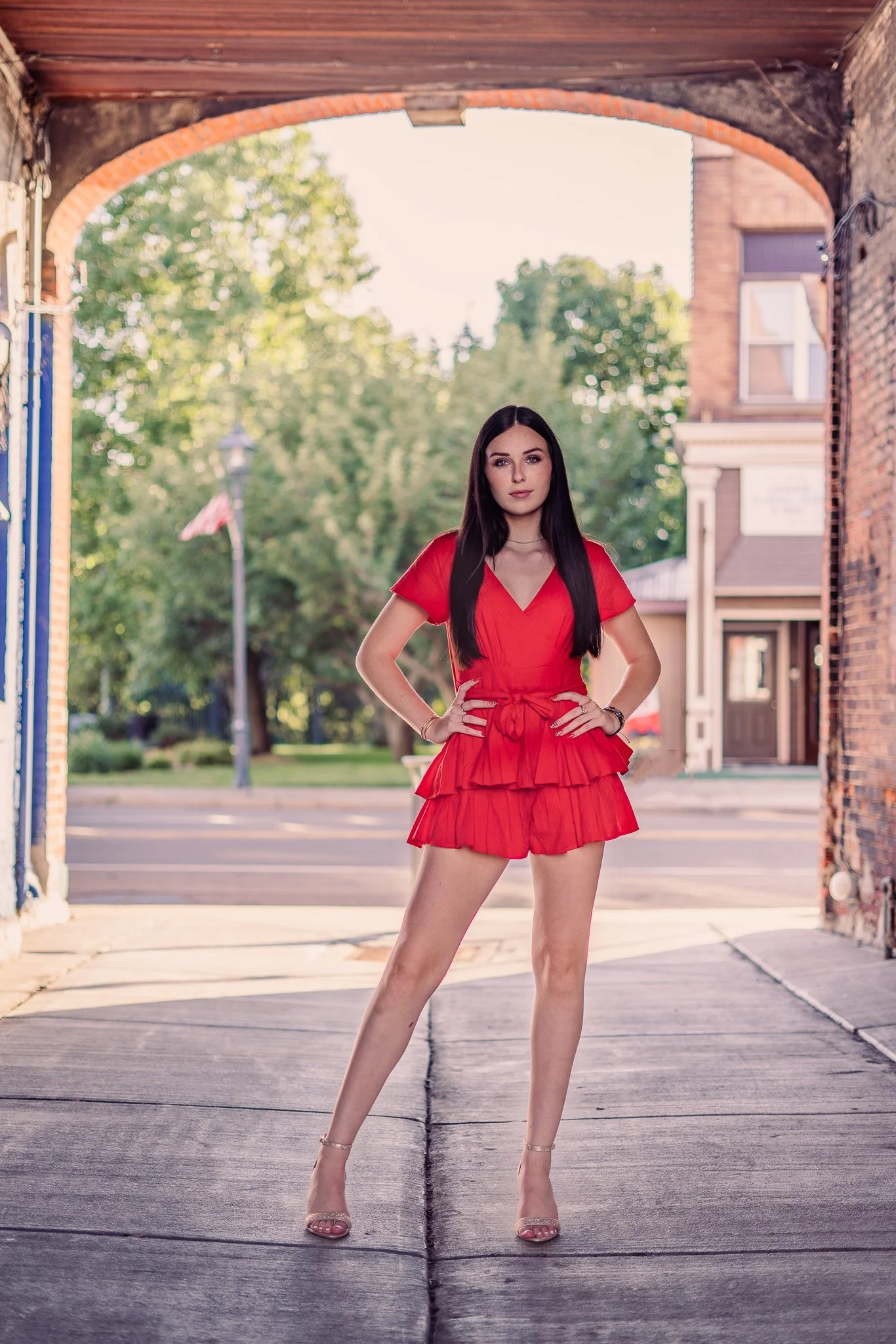 Young woman in red outfit standing confidently under an archway in an urban setting with trees and buildings in the background. Westfield, NY