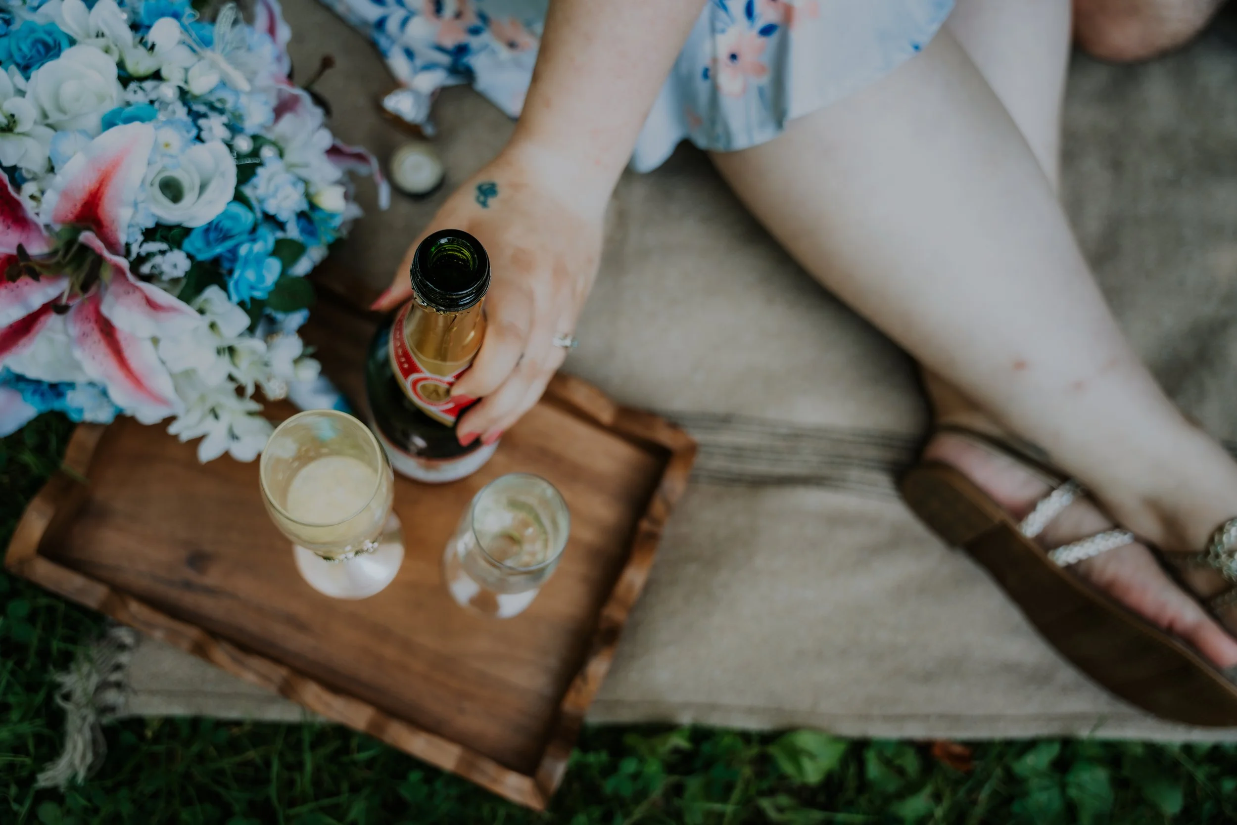 Person sitting outdoors on a beige mat, holding a champagne bottle, with a floral bouquet and glasses of champagne nearby. Fredonia, NY