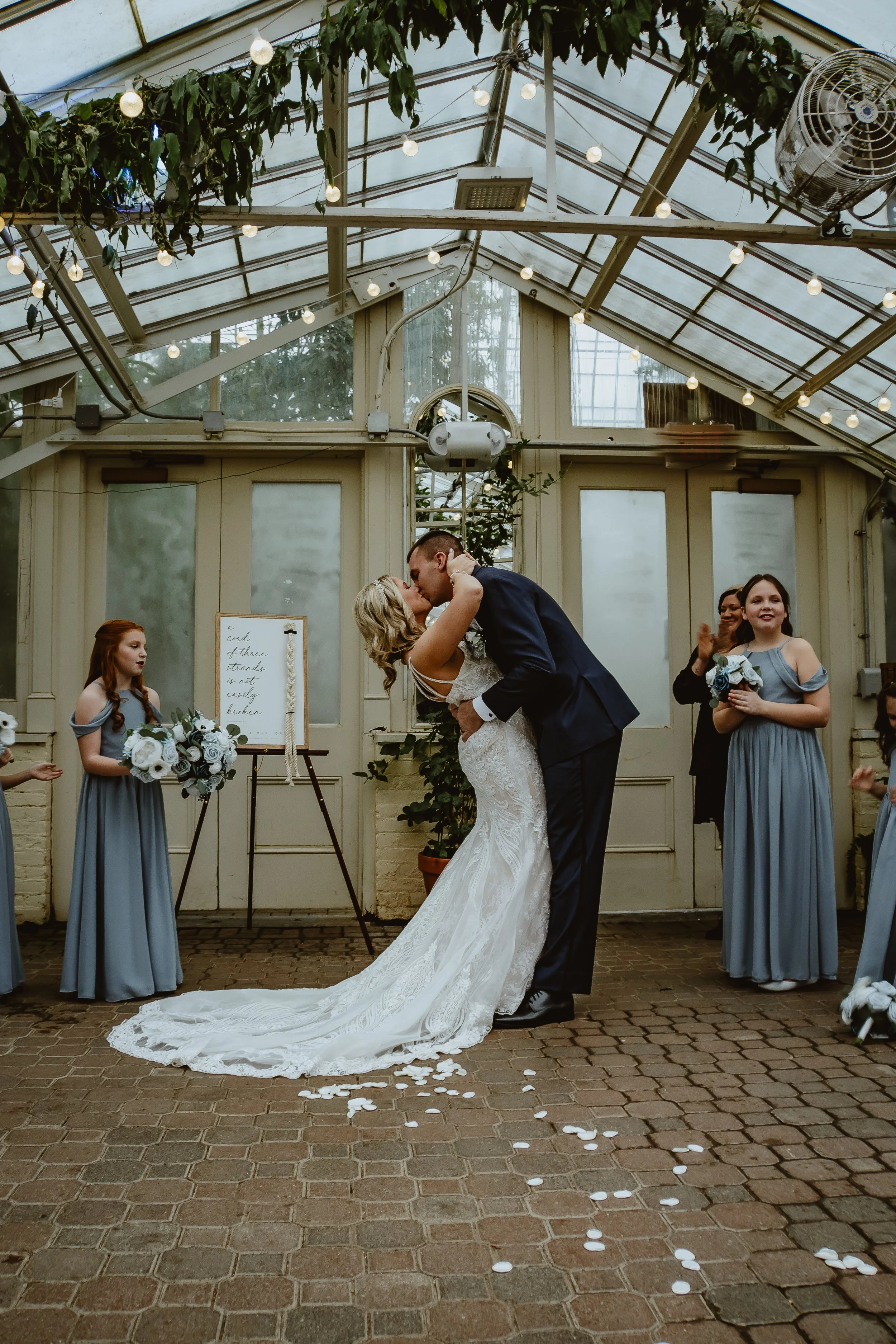 A bride and groom kiss during their wedding ceremony in a greenhouse, surrounded by bridesmaids holding bouquets, with flower petals on the floor and string lights overhead.