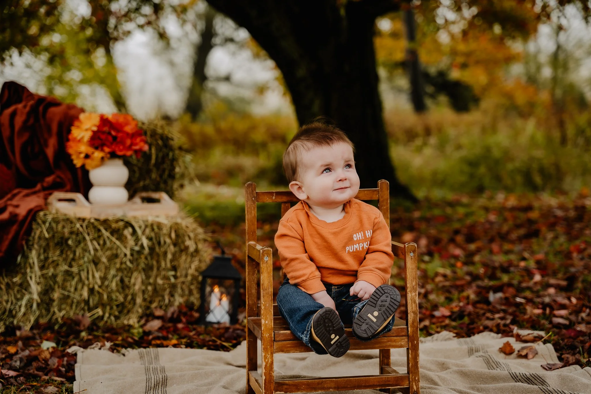 A young child sitting on a small wooden chair outdoors in autumn, surrounded by fallen leaves. The child is wearing an orange sweater and jeans, gazing upward. In the background, there are trees with fall foliage, a hay bale, a lantern, a white vase 