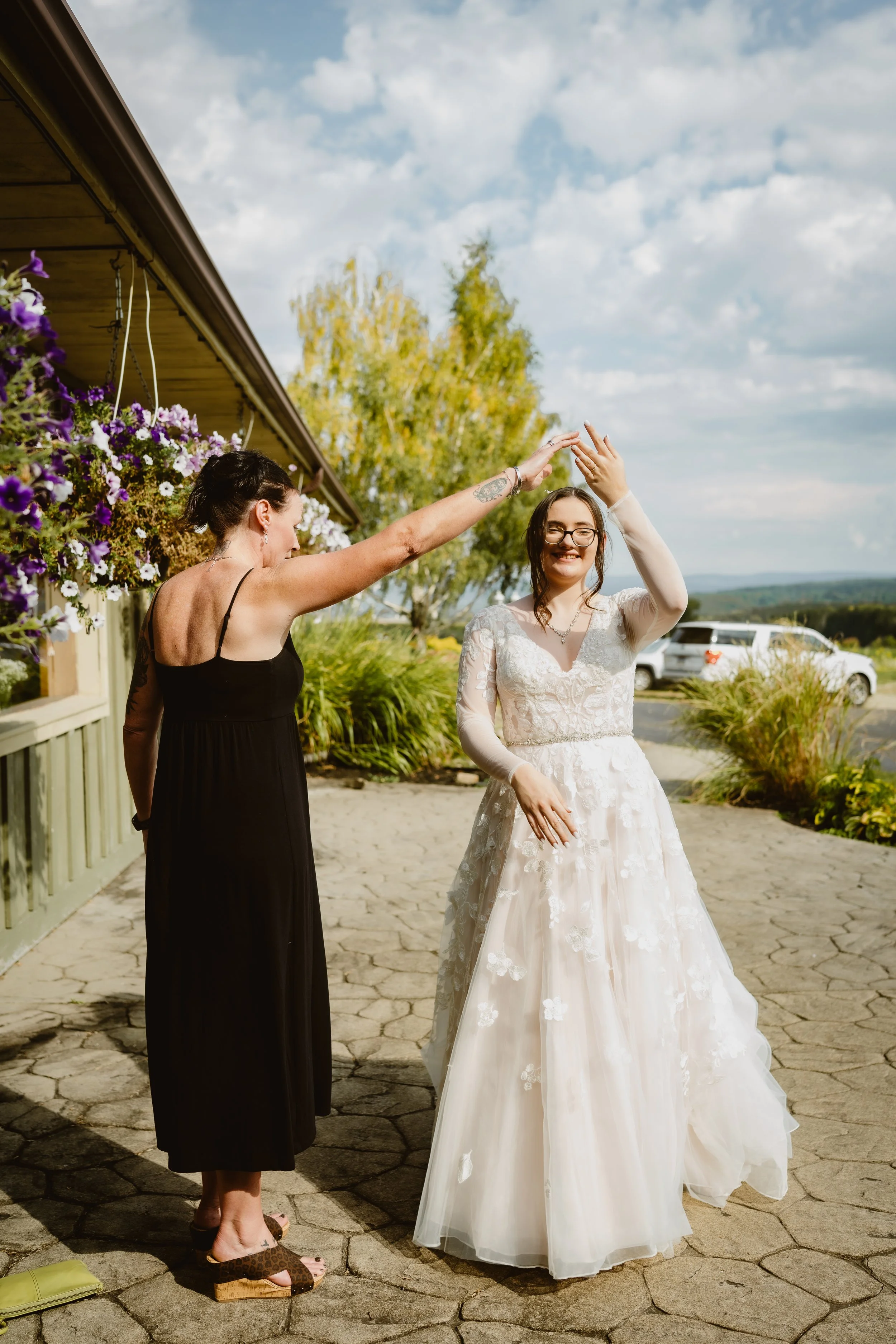 A woman in a wedding dress and a woman in a black dress dancing outdoors on a stone patio with purple and white flowers hanging from a building, trees, and a car in the background. The Grandview of Ellington, Ellington NY