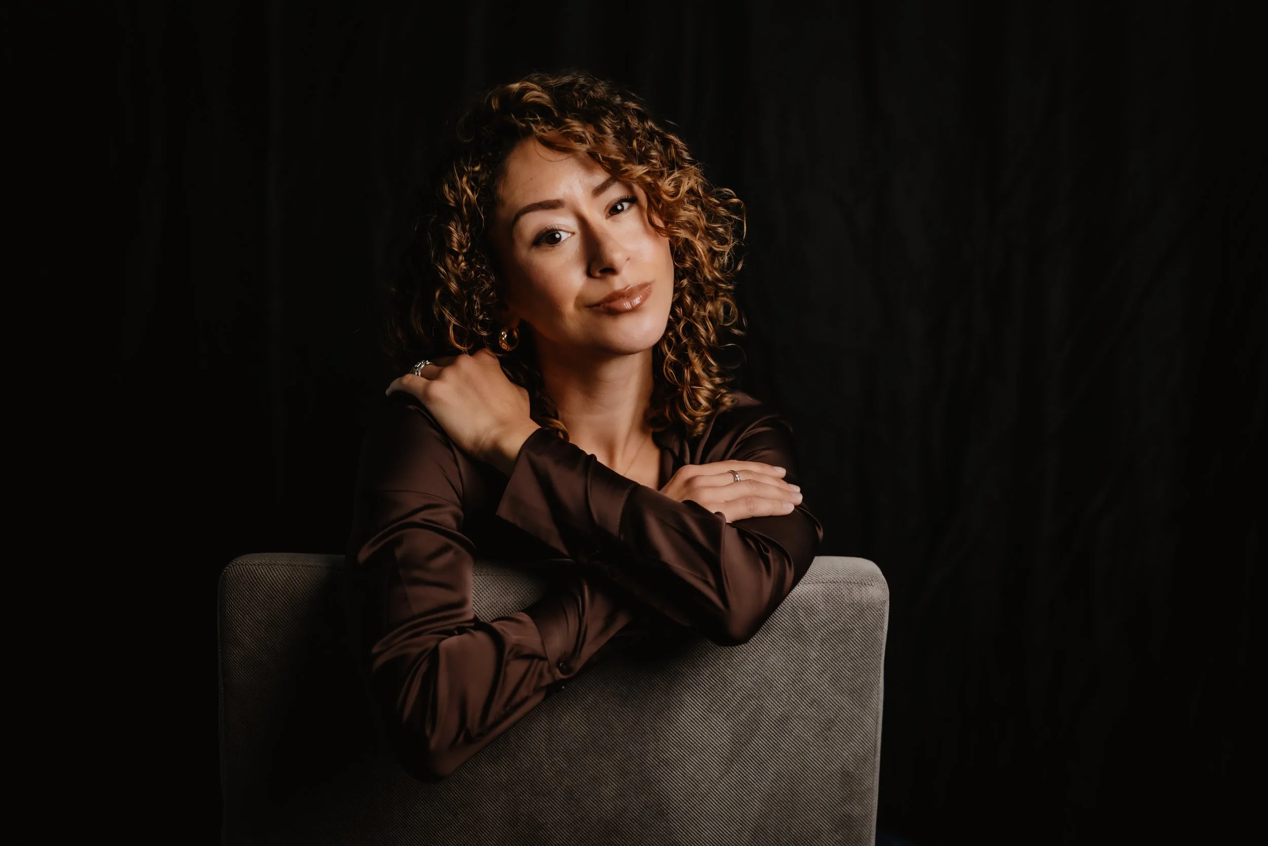 A woman with curly brown hair wearing a shiny dark brown blouse, seated behind a gray chair, posing against a black background.