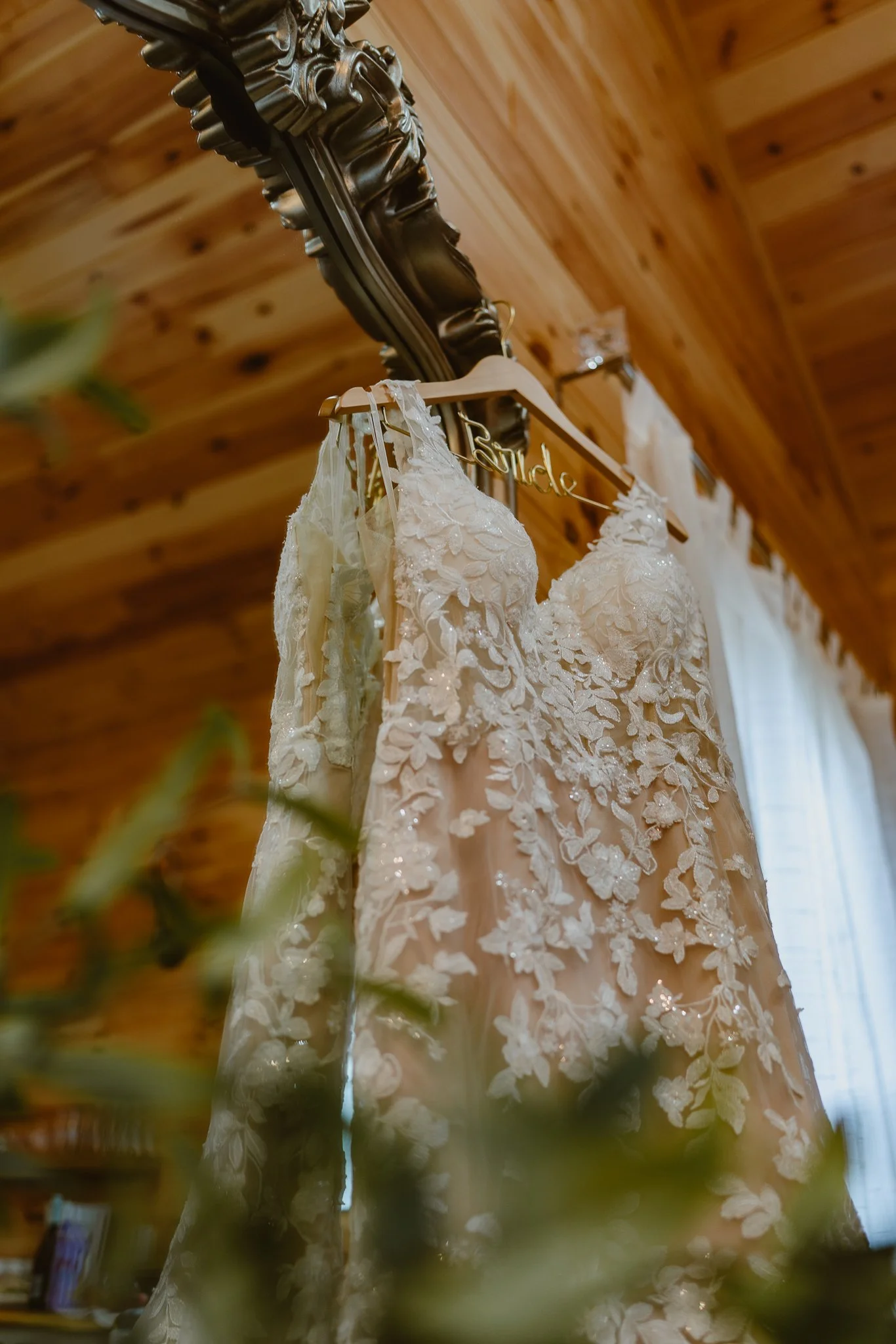 A wedding dress hangs on a hanger from a decorative wooden frame against a wooden ceiling. The dress is detailed with floral lace embroidery.