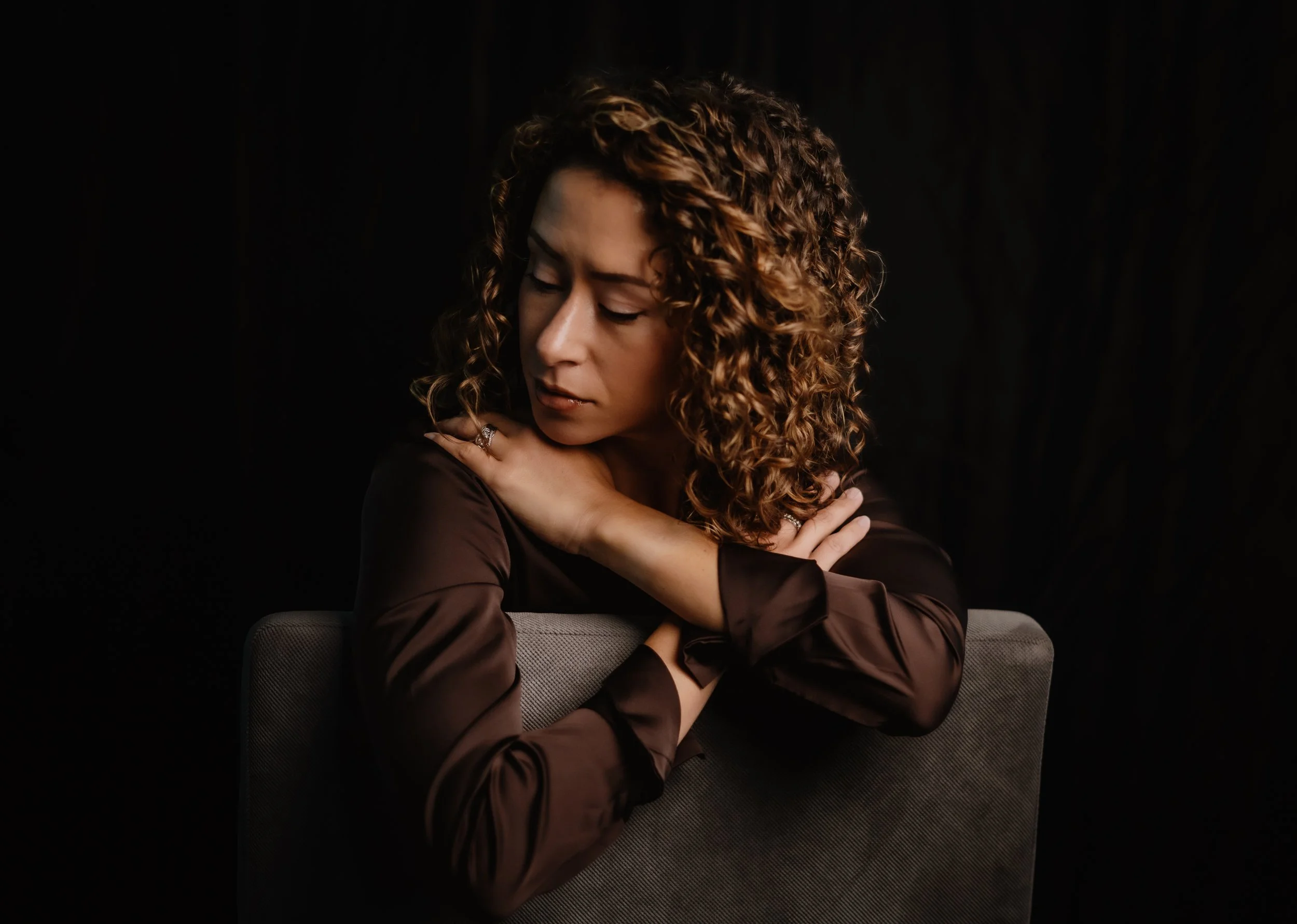 A woman with curly hair wearing a satin brown blouse, resting her arms on a grey chair, with a black background, and a contemplative expression on her face.