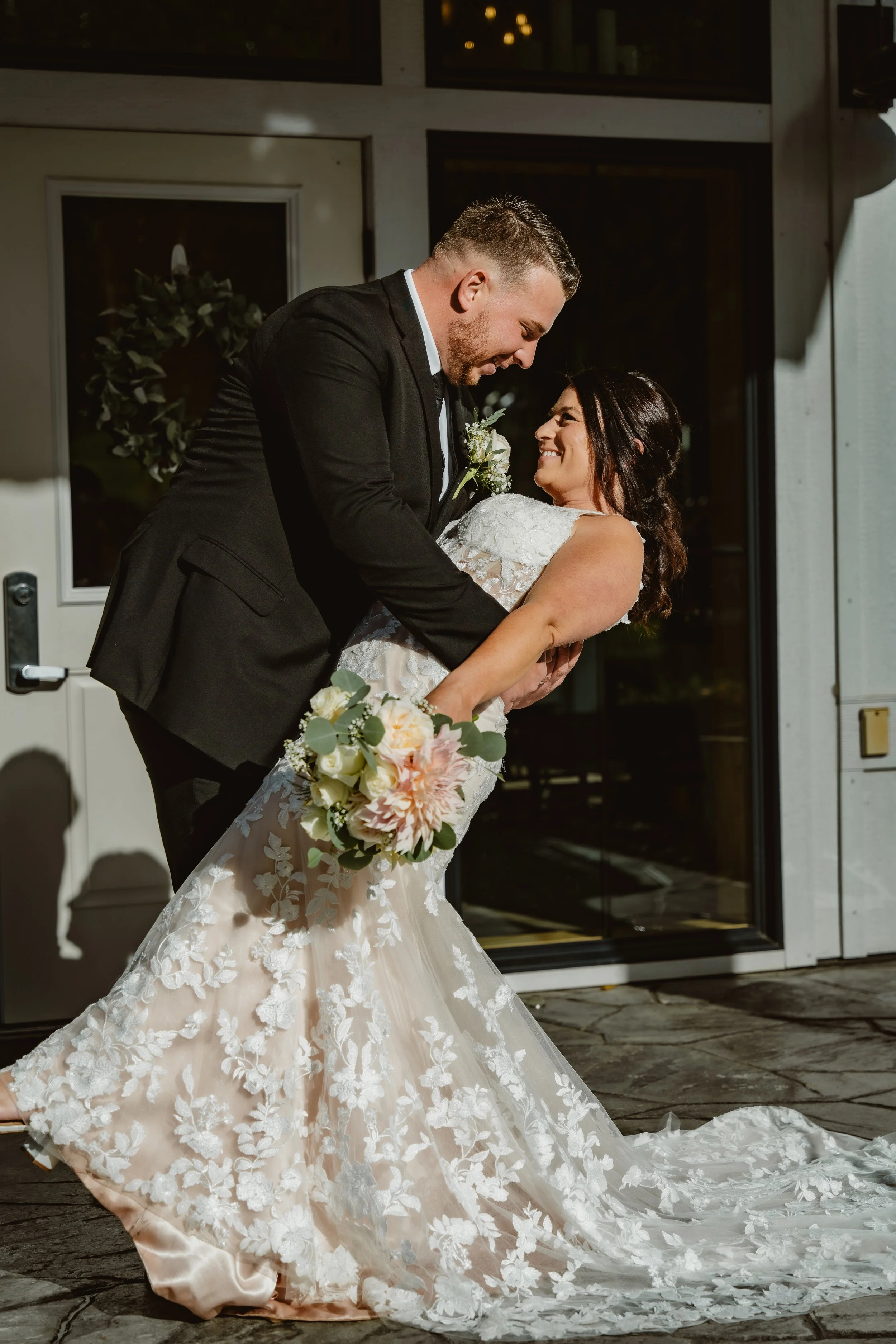 A bride and groom share a dance outside, smiling at each other. The bride is holding a bouquet of flowers, wearing a lace wedding gown, and the groom is wearing a black suit. The Barn at Farrington Hollow, Cherry Creek, NY