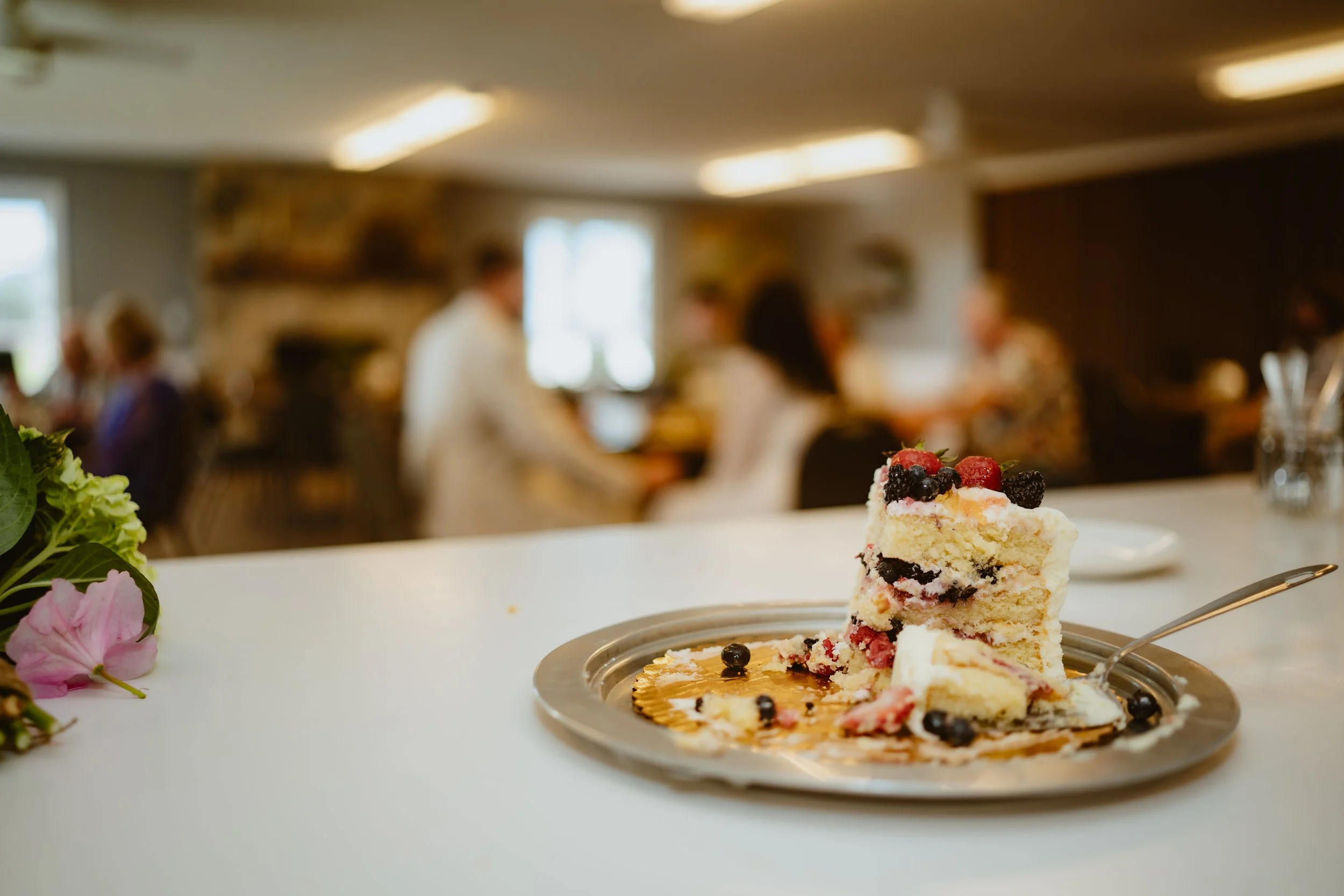 A partially eaten slice of berry cake on a silver plate with a fork, on a white counter, with a blurred background of people and a cozy indoor setting.