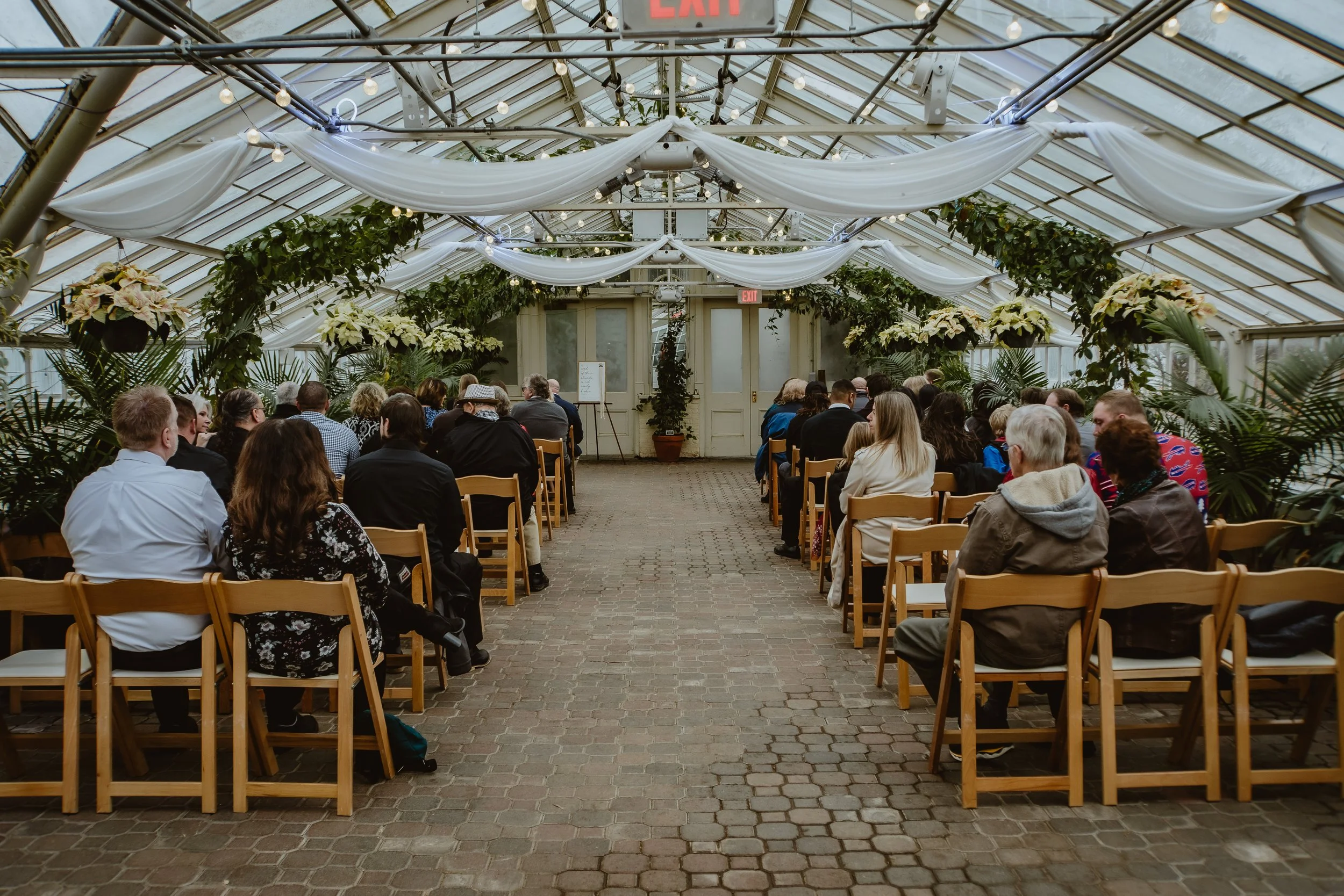 People seated in a greenhouse decorated with white fabric and green plants, attending a wedding or event.