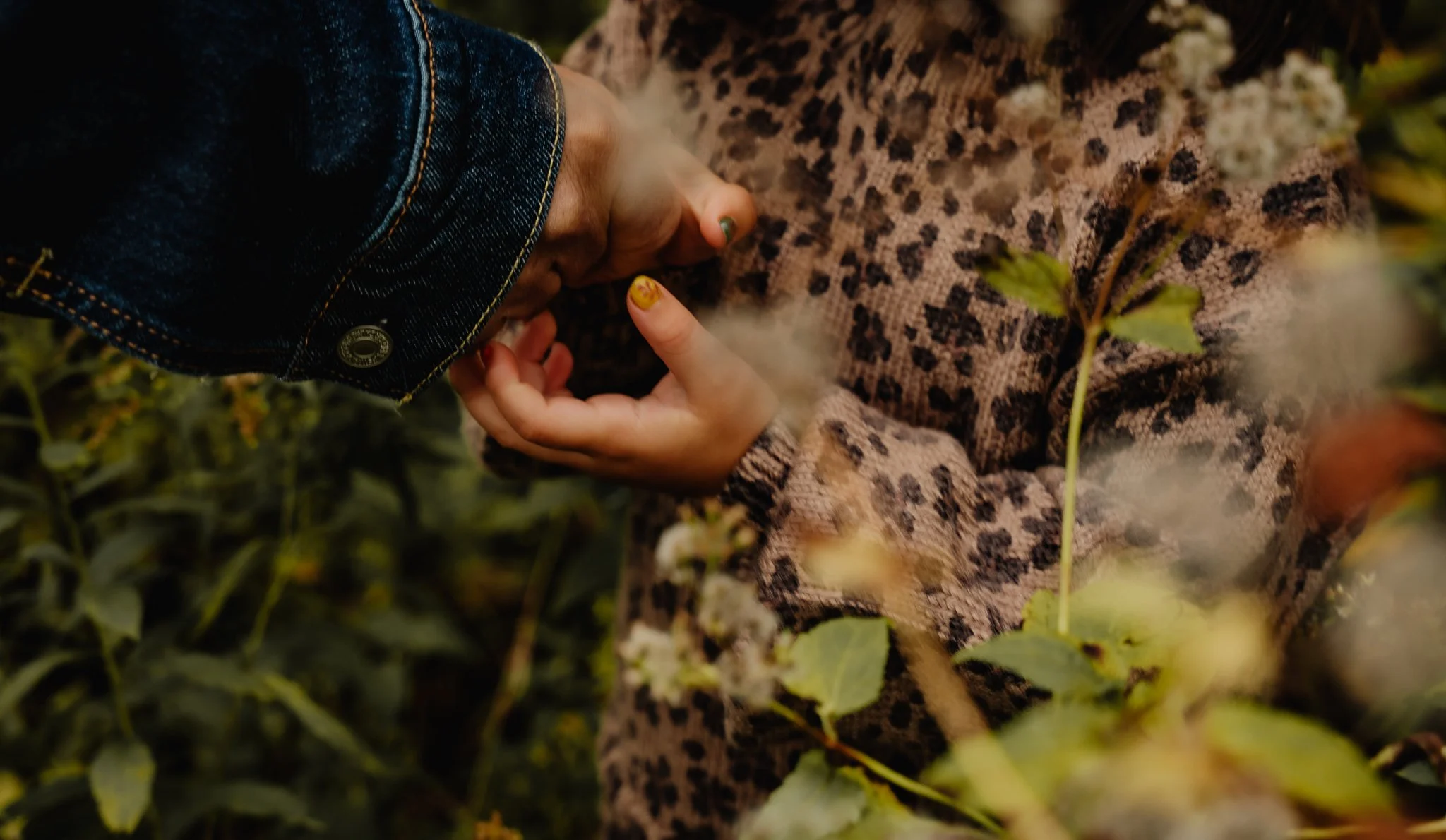 Person lying among plants, wearing a leopard print coat and denim jacket, with their eyes closed.