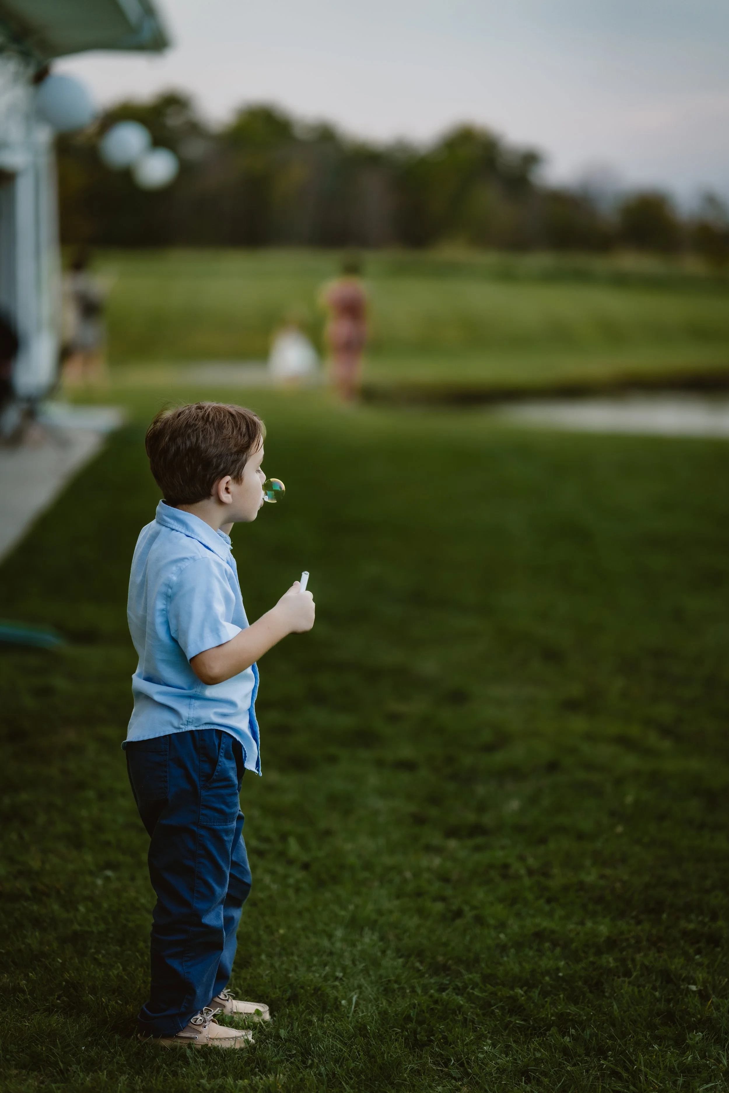 A young boy in a light blue shirt and dark blue pants blowing bubbles outdoors on a grassy area during daytime. The Grandview of Ellington, Ellington NY
