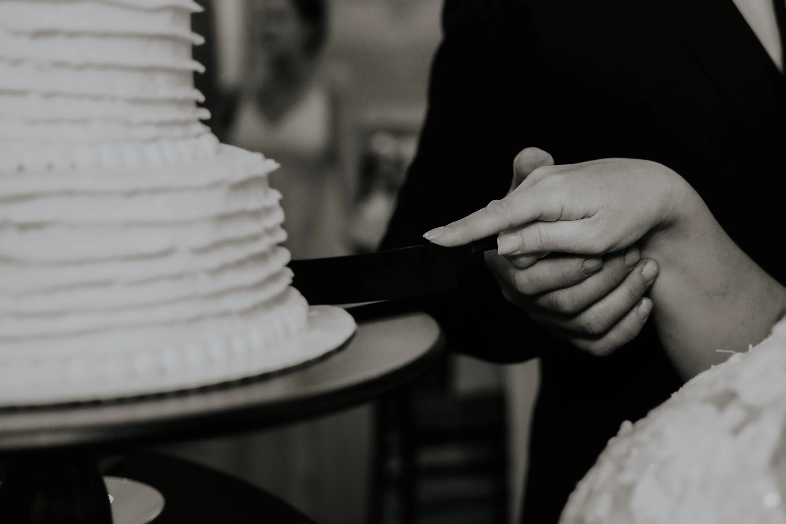 Close-up of a two people's hands slicing a layered wedding cake with white frosting, black background. Farrington Hollow, Cherry Creek, NY