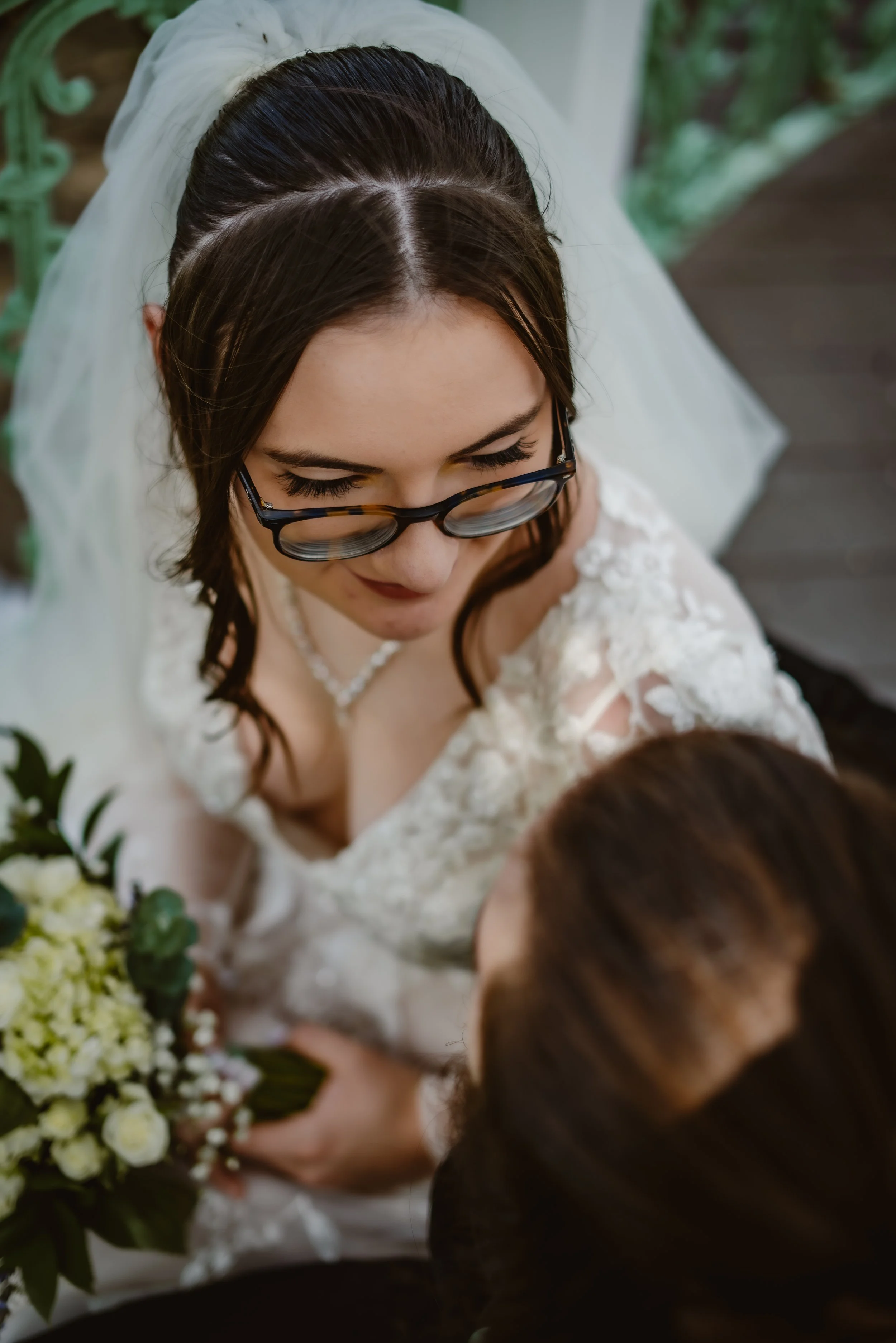 A woman in a white wedding dress and glasses holds a bouquet of white flowers and gazes down at another person, possibly a bride, in a close, intimate moment.