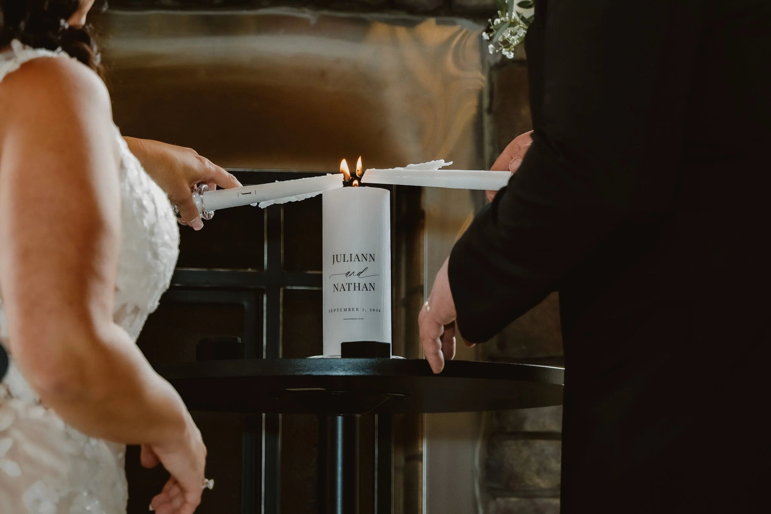 A wedding candle-lighting ceremony, with two large white candles labeled with the names "Juliann and Nathan," and the date "September 7, 2024," being lit by the couple. The Barn at Farrington Hollow, Cherry Creek, NY