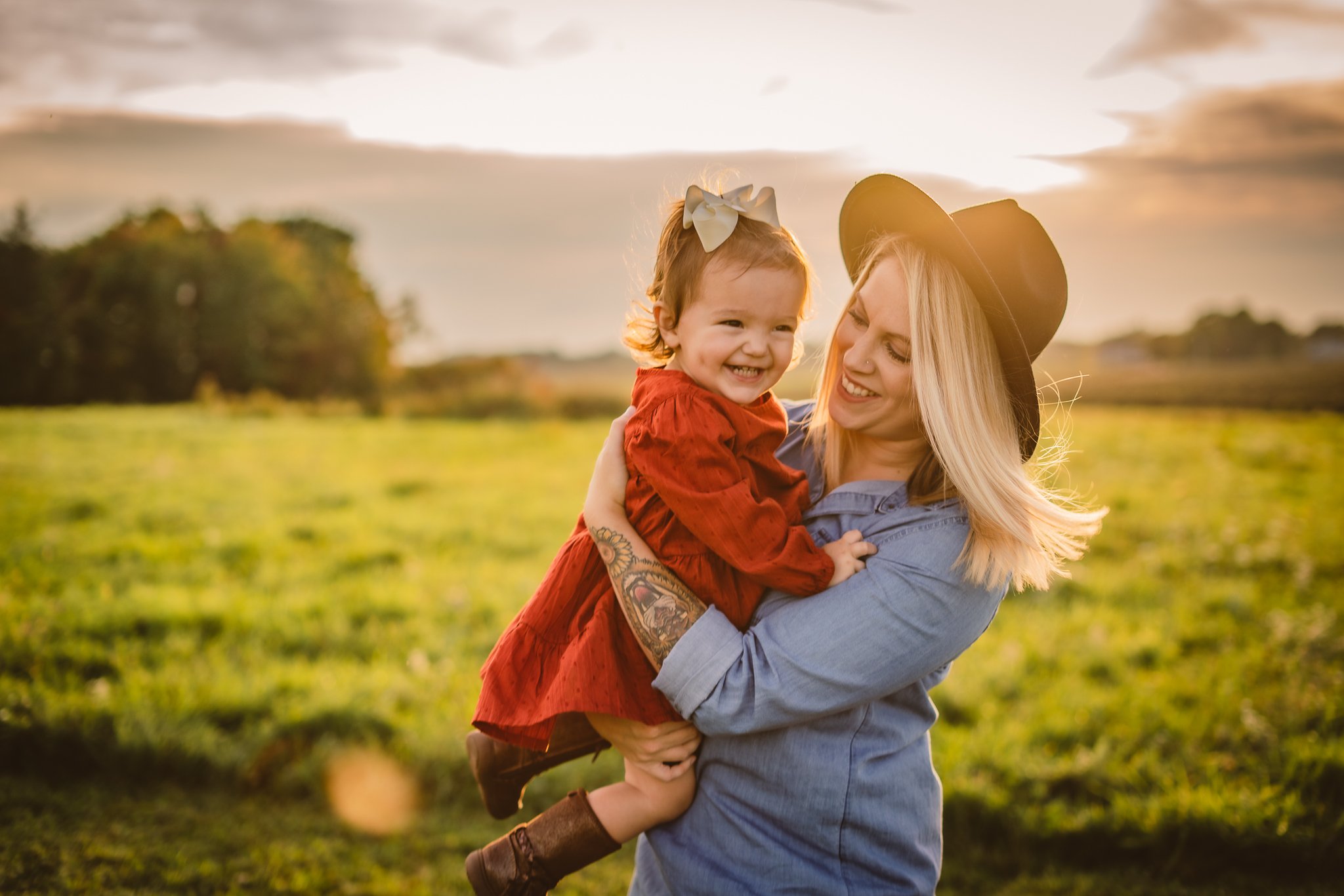 A woman with blonde hair in a denim shirt and black hat holding a young girl in a red dress with a white bow in her hair outdoors during sunset, smiling and looking at each other.