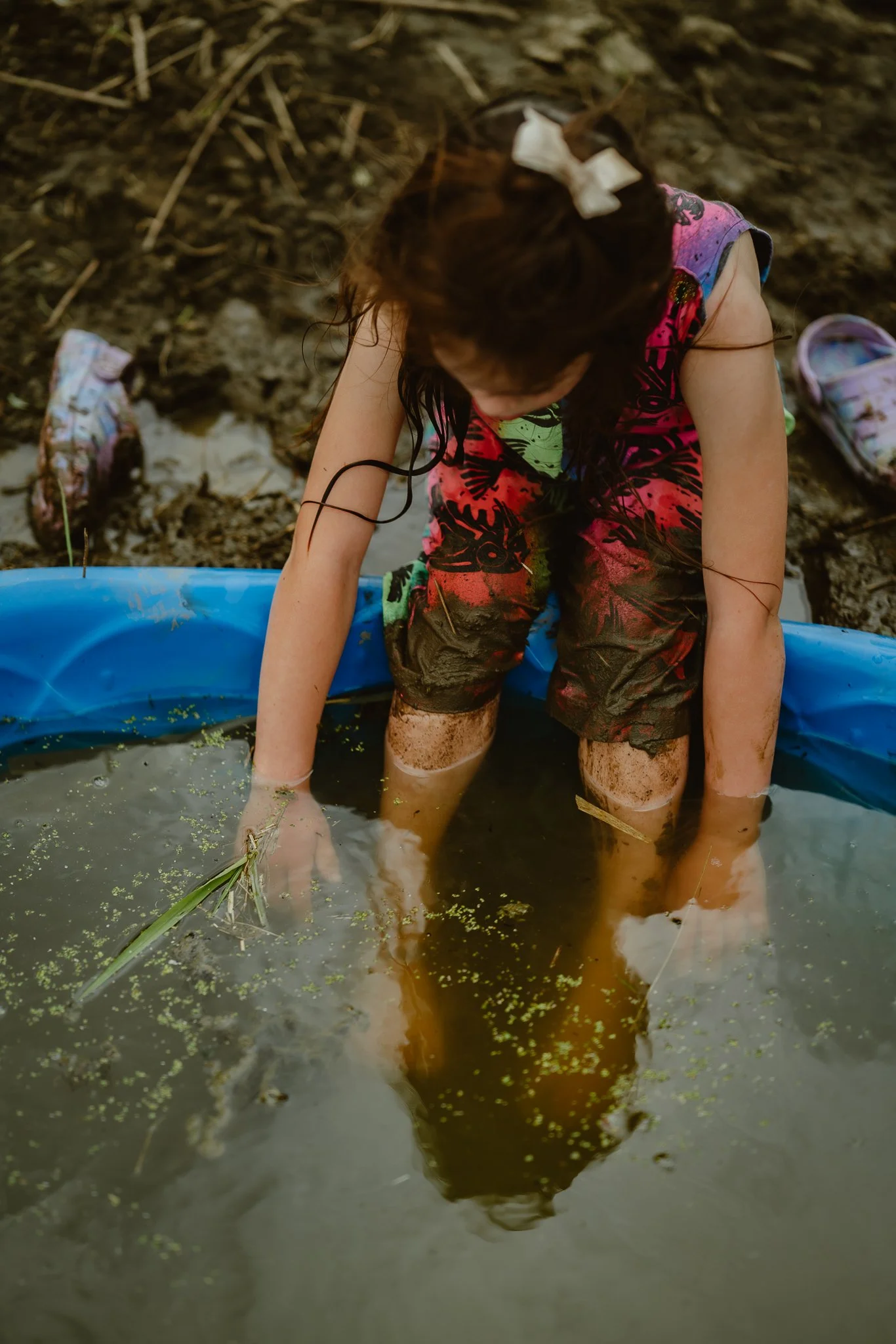 A young girl playing in a muddy pond, sitting with her legs in the water, wearing colorful clothes and a white bow in her hair, with shoes nearby on a muddy outdoor setting.