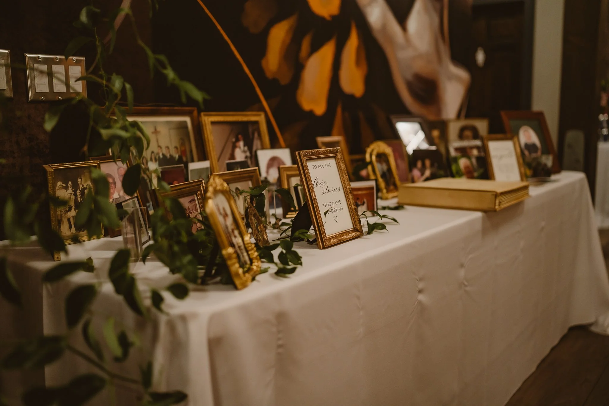 A display table with numerous framed photographs and a handwritten note, decorated with greenery, in a warmly lit indoor setting.