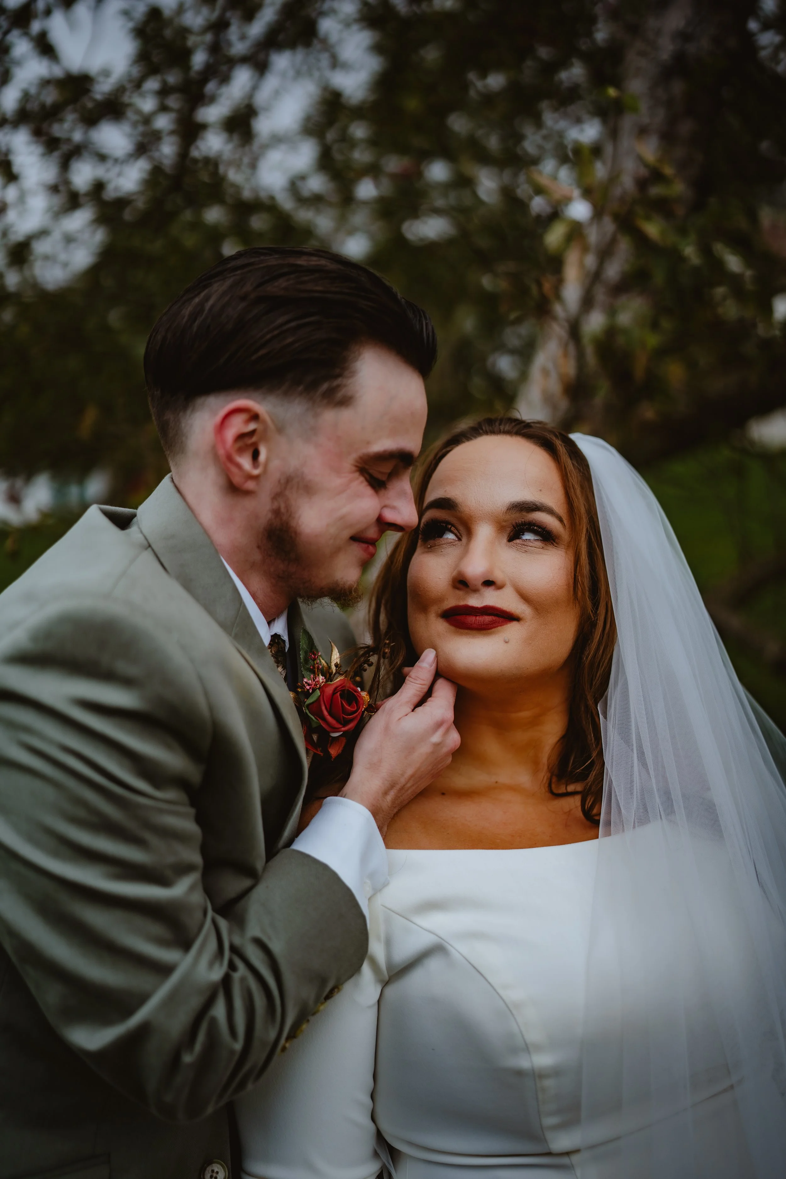 A groom and bride embracing outdoors, with trees and sky in the background. The groom, wearing a light gray suit with a boutonniere, gently touches the bride's chin, who is wearing a white wedding dress and veil. They are sharing an intimate moment, 