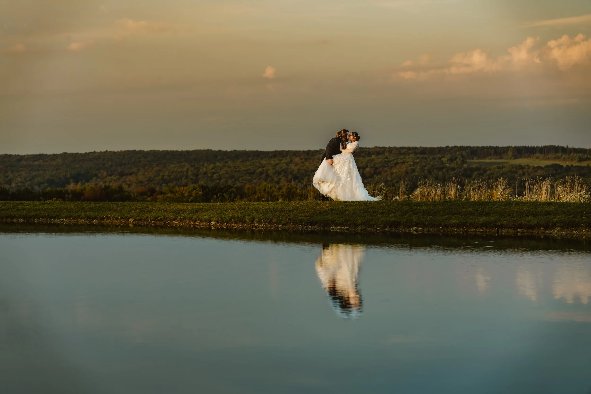 A bride and groom kissing outdoors near a body of water during sunset, with a scenic landscape in the background and their reflection visible in the water. The Grandview of Ellington, Ellington NY