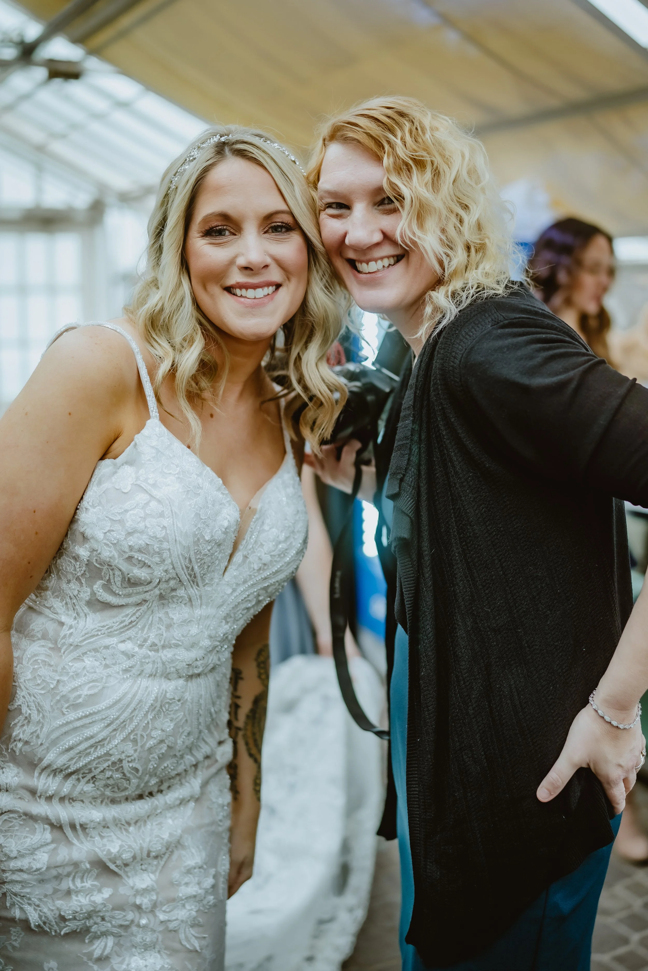 Two women smile and pose for a photo, one wearing a wedding dress and the other with curly blonde hair and casual clothing, in a well-lit indoor setting. Buffalo Botanical Garden
