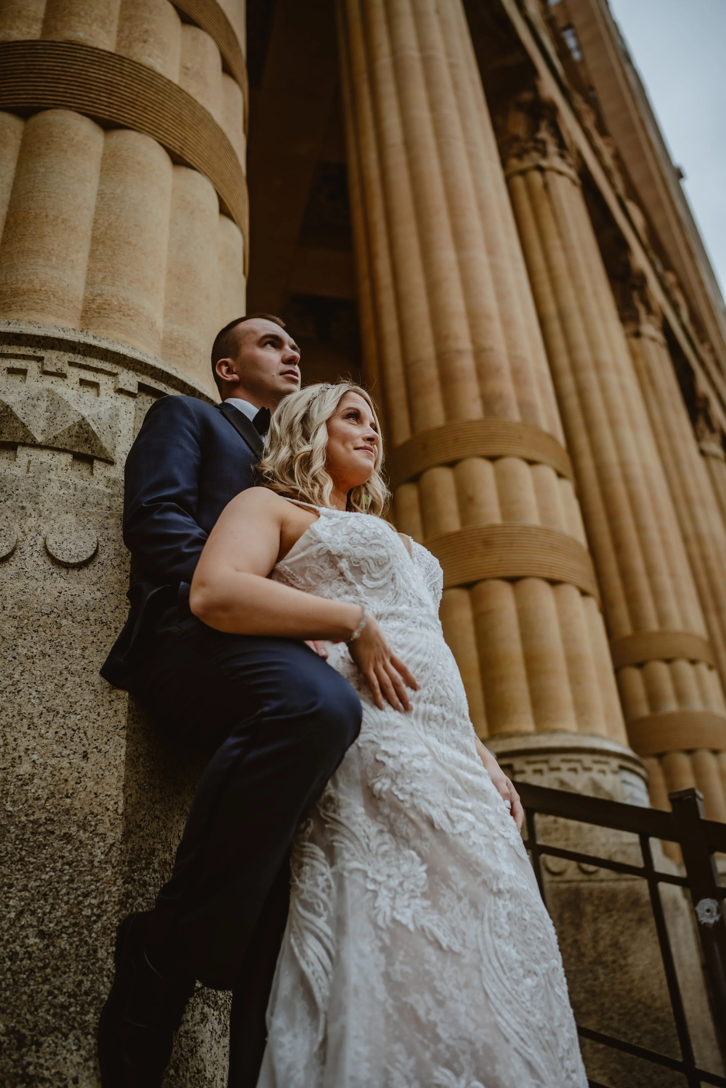 A newlywed couple stands outside a historic building with large columns, looking upwards. The bride is in a white wedding dress, and the groom is in a dark tuxedo. Buffalo NY