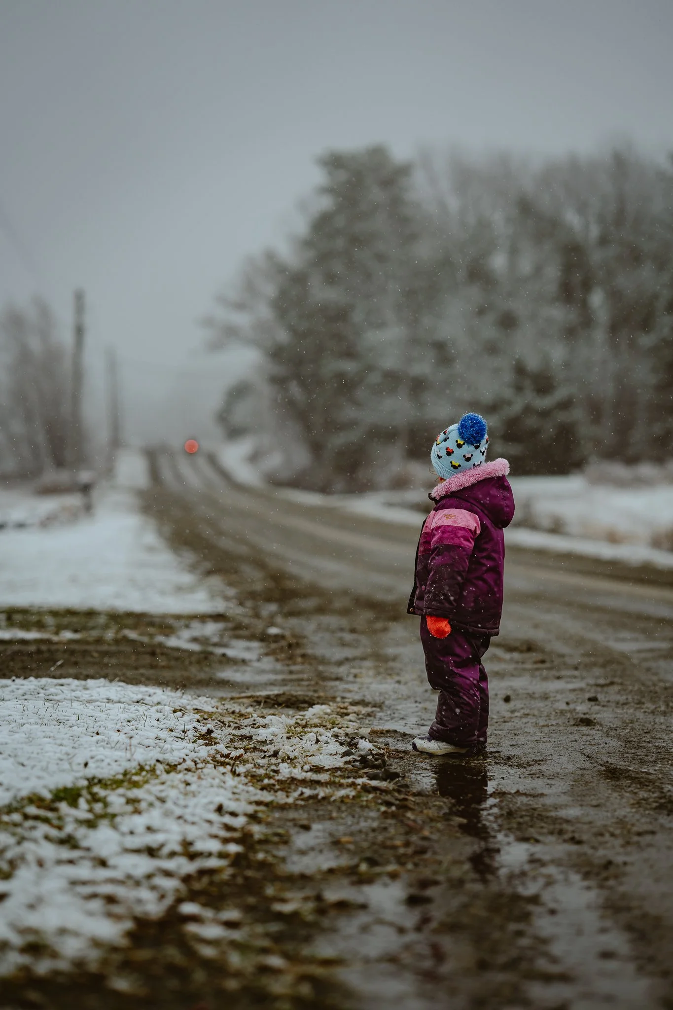 A young child dressed in a purple winter coat, snow pants, orange gloves, and a colorful knit hat standing on a snowy, muddy road during snowfall, looking into the distance with trees and a train track in the background.