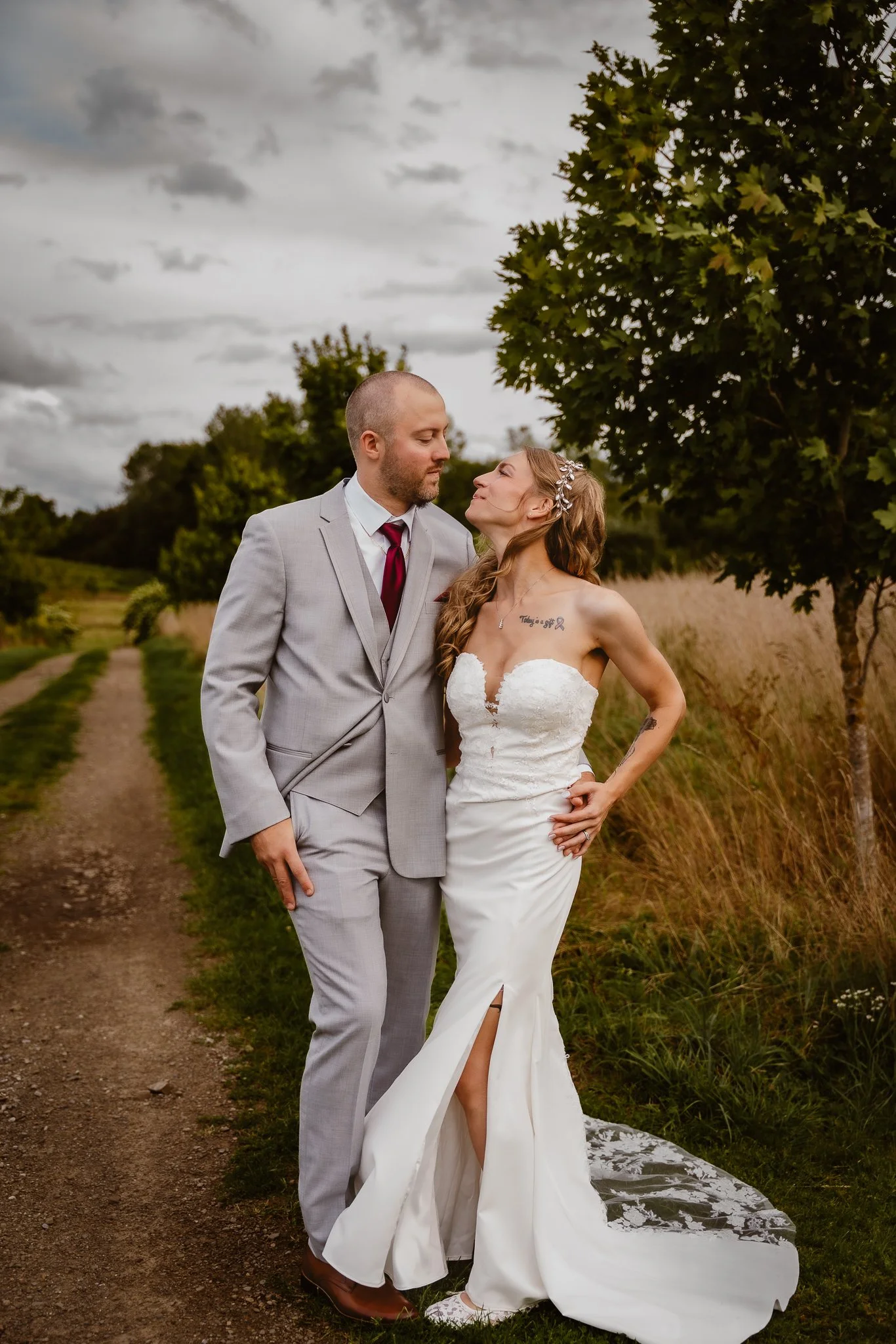 A newlywed couple stands on a dirt path in a field with tall grass and trees, gazing into each other's eyes on a cloudy day. CHQ barn, Mayville NY