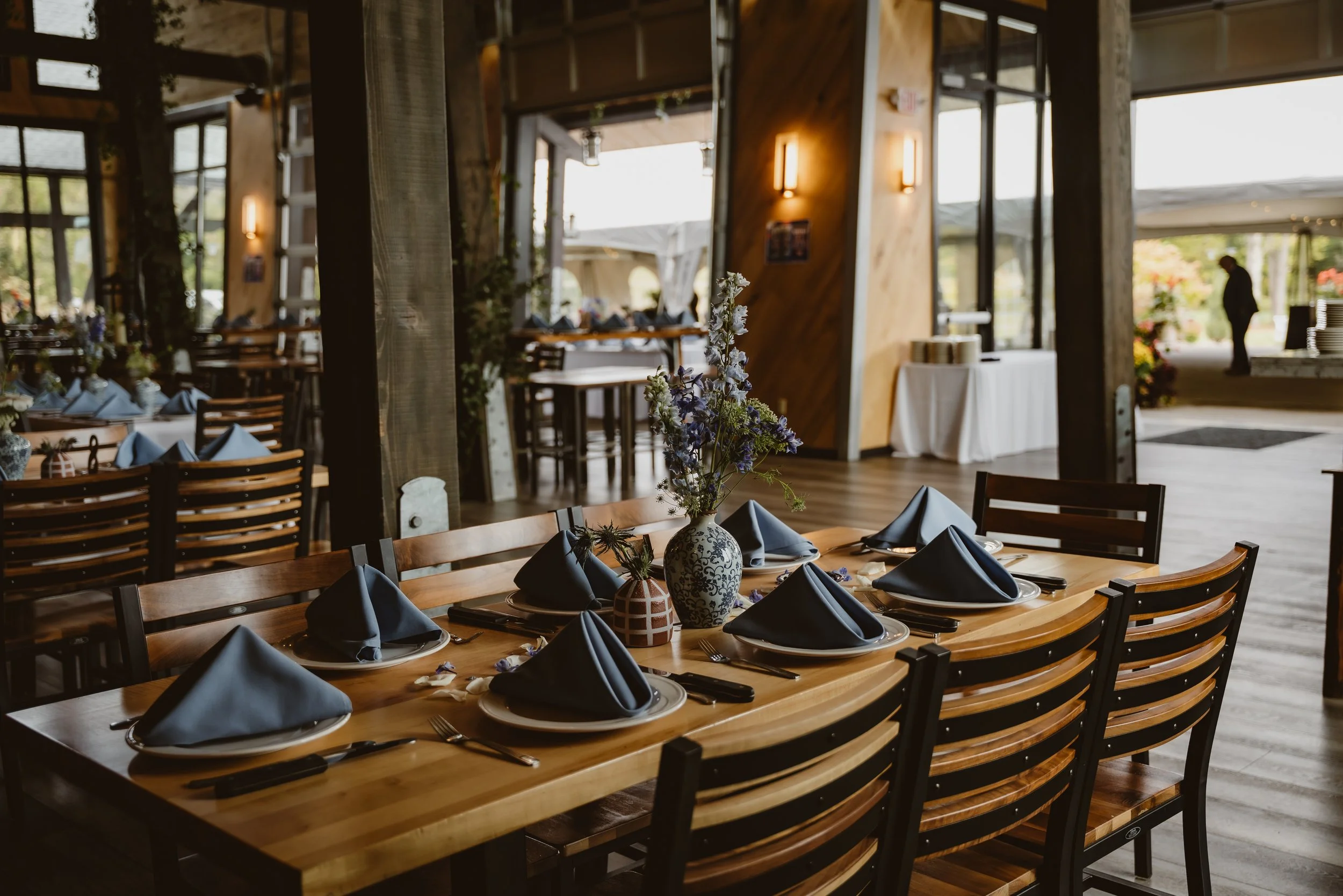 A restaurant with a wooden table set for a meal, featuring dark napkins, plates, and cutlery, and decorated with a floral centerpiece in a vase. The interior has wooden accents, large windows, and natural light. Ellicotville Brewing Company EBC